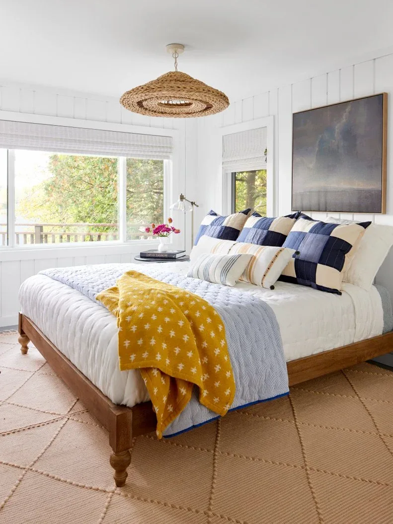 White wall boards surround a timber bed with checked blue cushions, a yellow throw and a woven ceiling light.