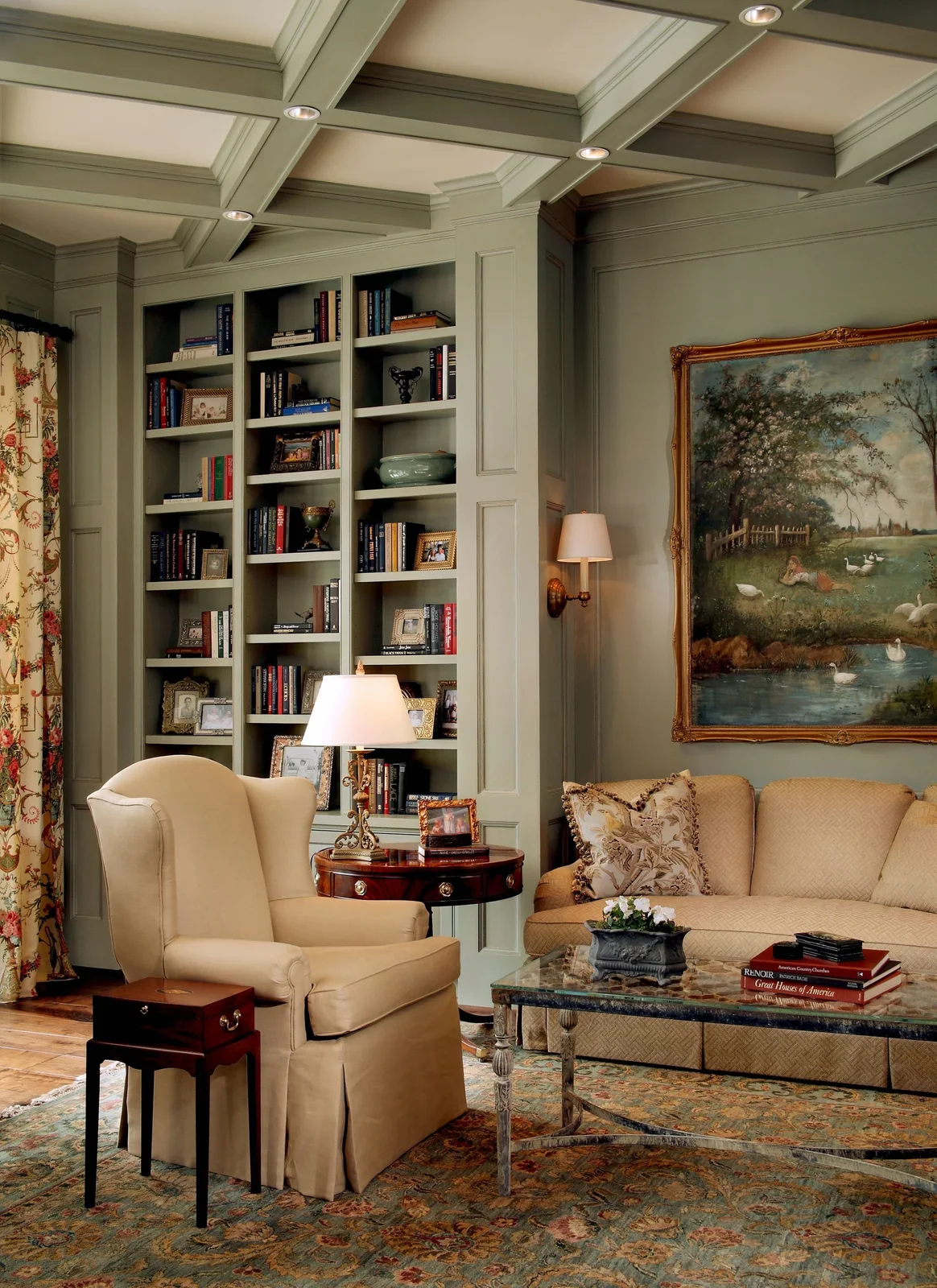 Pale paneled walls and bookshelves surround a cream armchair and sofa beside a table lamp, framed landscape and patterned rug