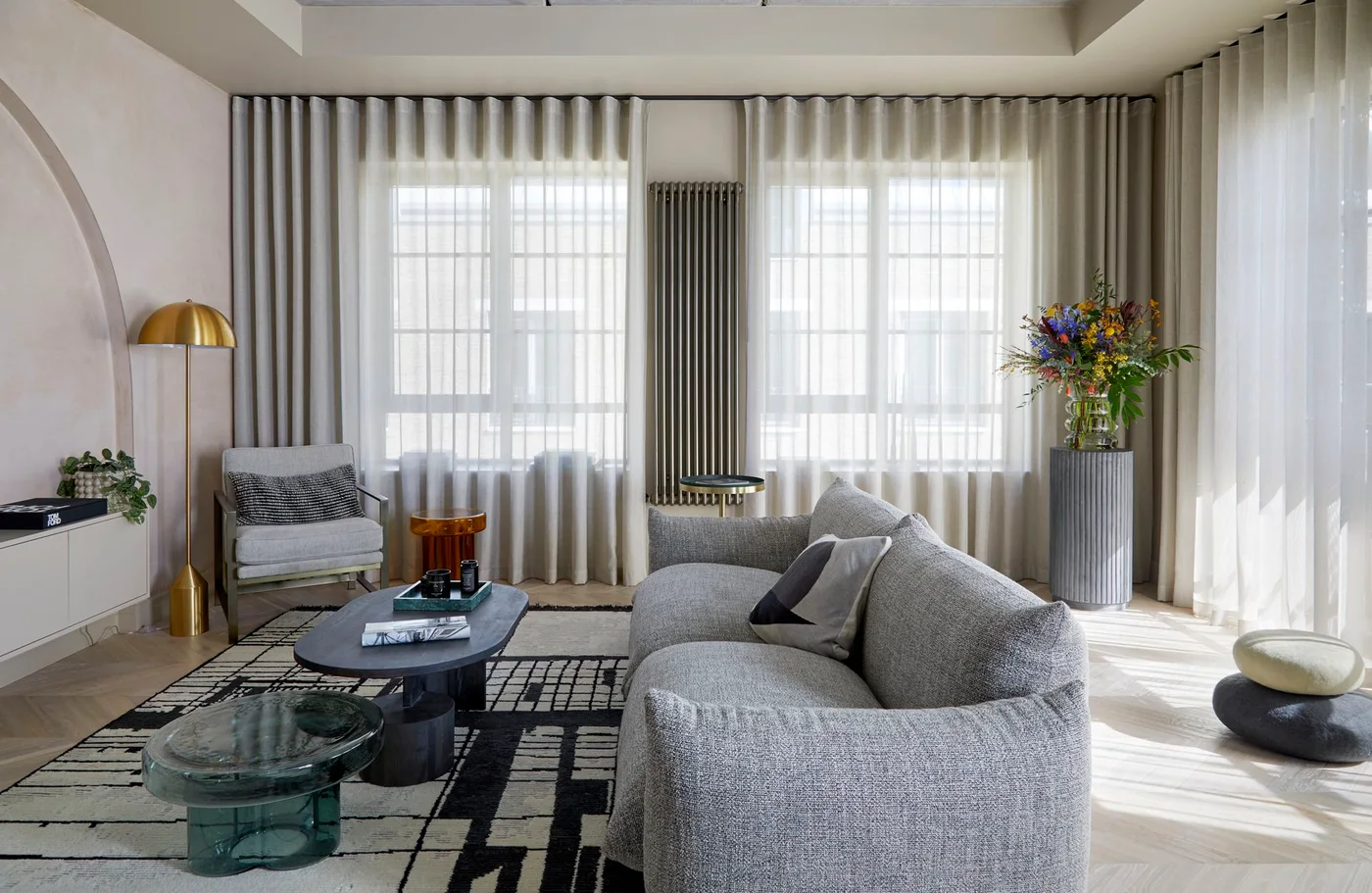 A gray curved sofa sits beside a pale chair, dark round tables and a glass stool above a patterned rug with long pale curtains behind