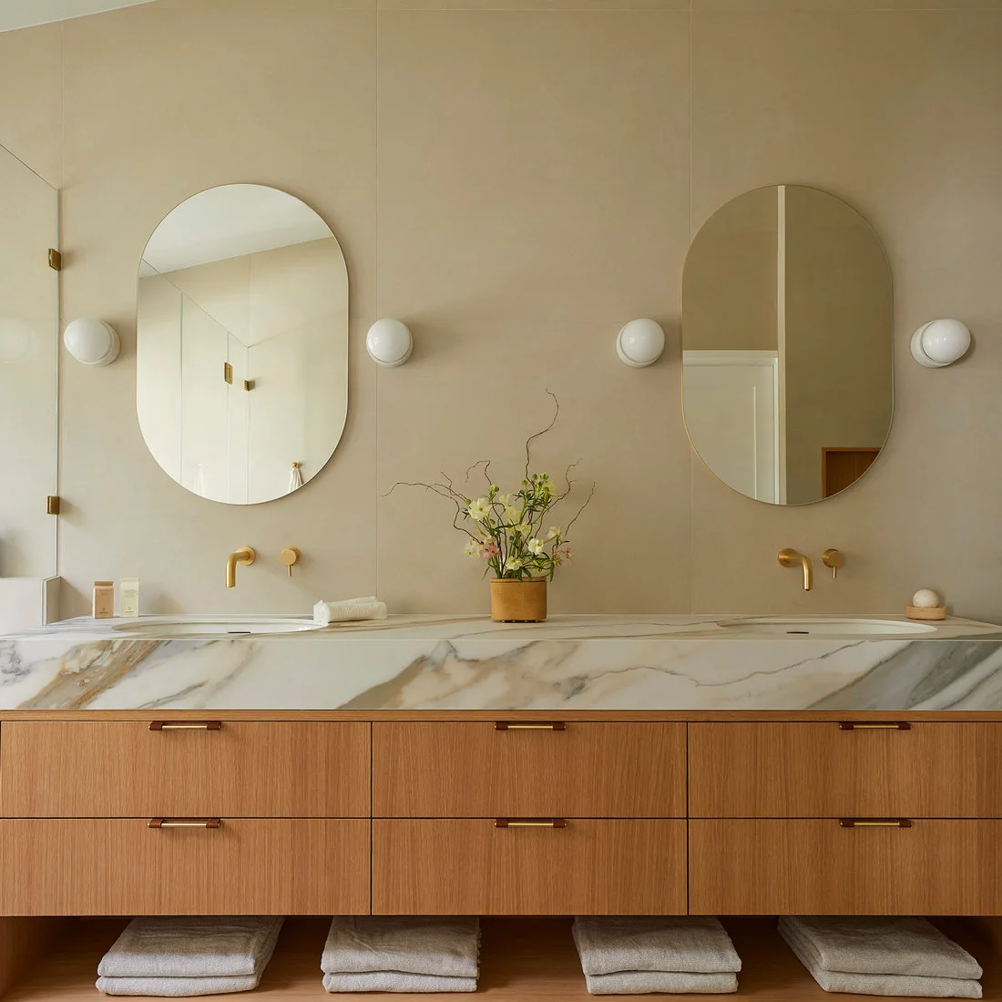 A warm wood double vanity has a pale veined counter, two long oval mirrors, warm-metal wall taps, white globe lights and open towel shelves below.