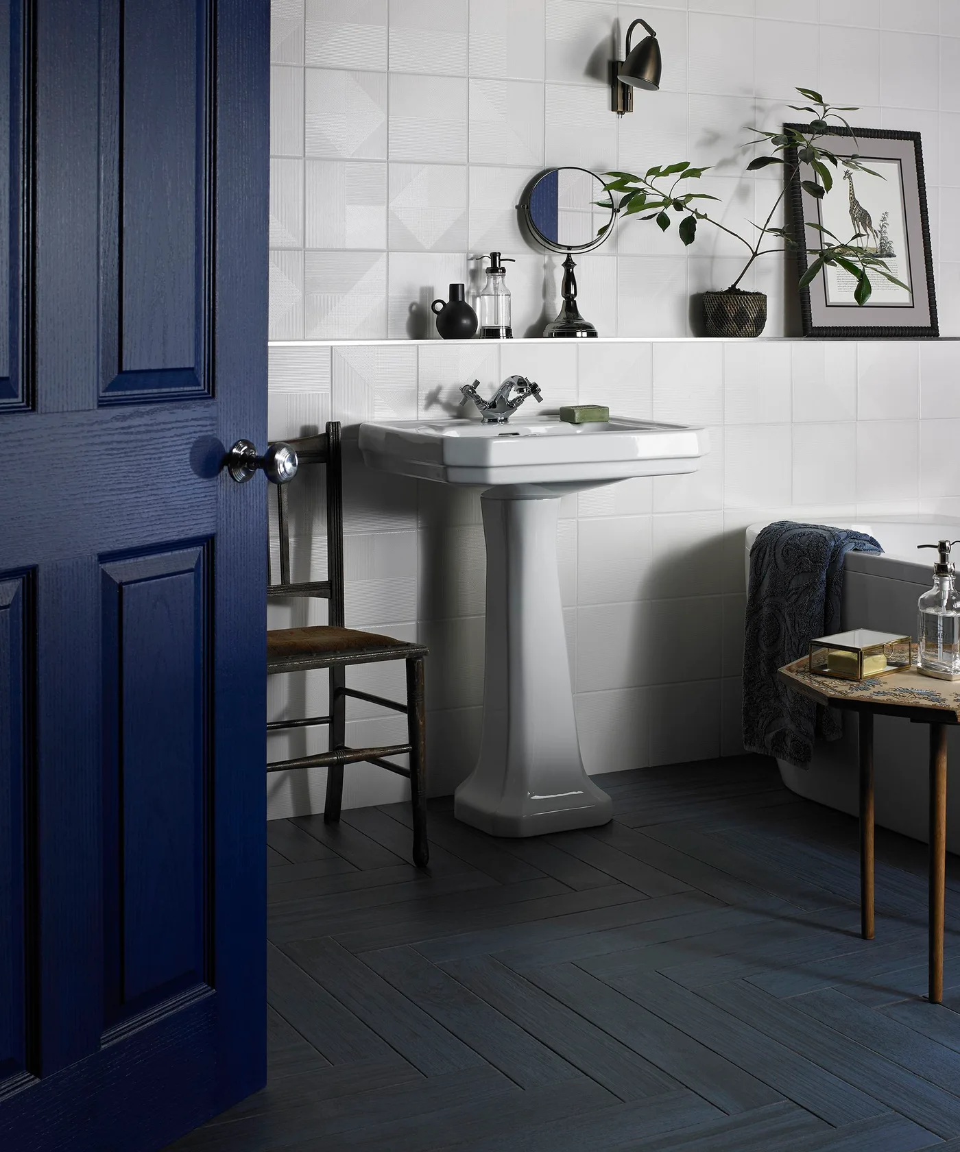 A navy paneled door opens into a bathroom with white wall tiles, a pedestal sink and a dark floor
