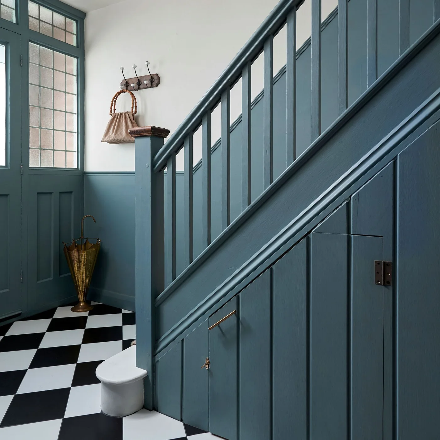 Blue-green entry door and stair balustrade with paneled storage doors below, checked floor, wall hooks and umbrella container