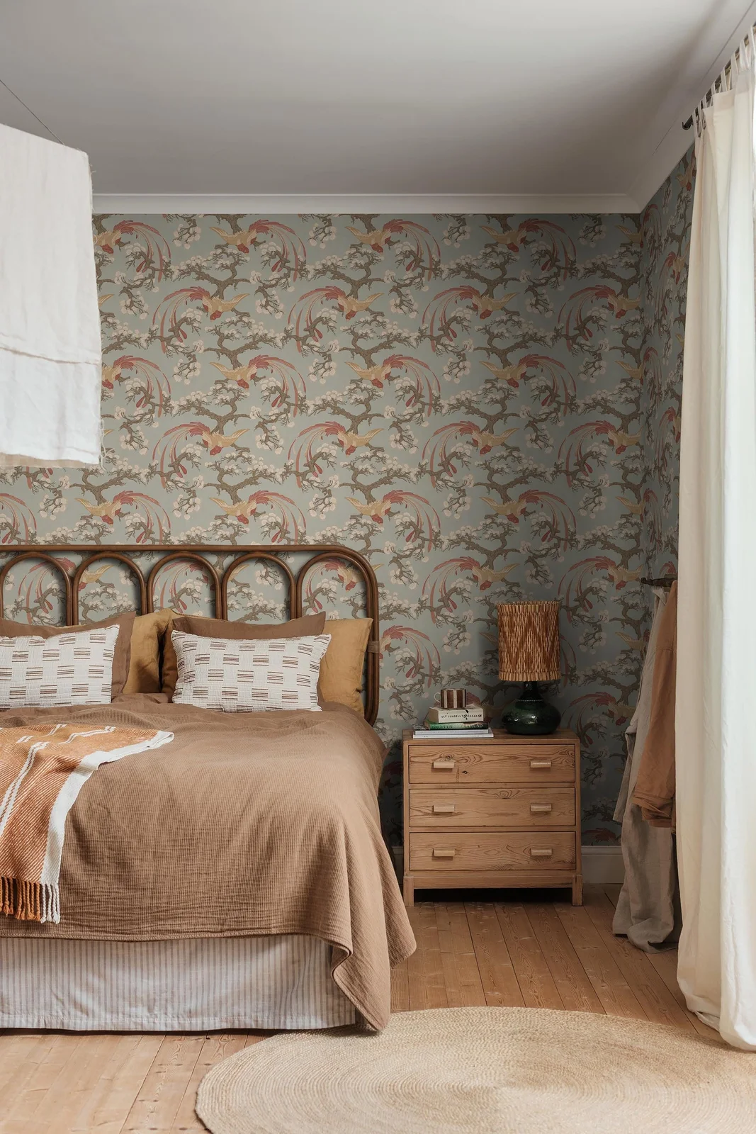 Muted bird-and-branch wallpaper surrounds a cane headboard, warm brown bedding, wood bedside drawers and a dark lamp with a woven shade.