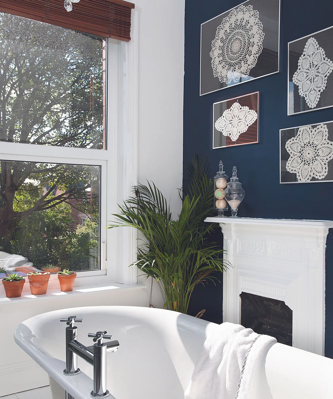 A navy wall displays framed white lace-like pieces above a white fireplace beside a freestanding bath