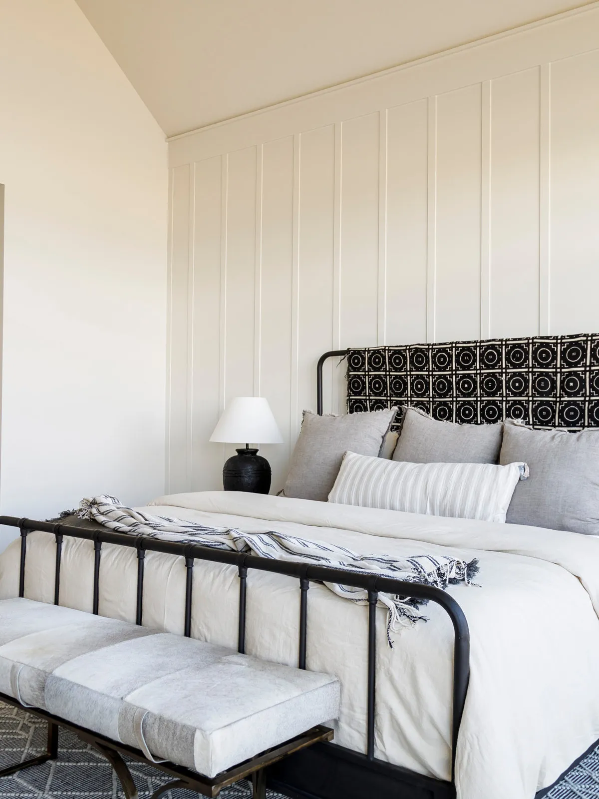 A black metal bed stands against pale vertical paneling with a patterned headboard cushion behind the pillows.