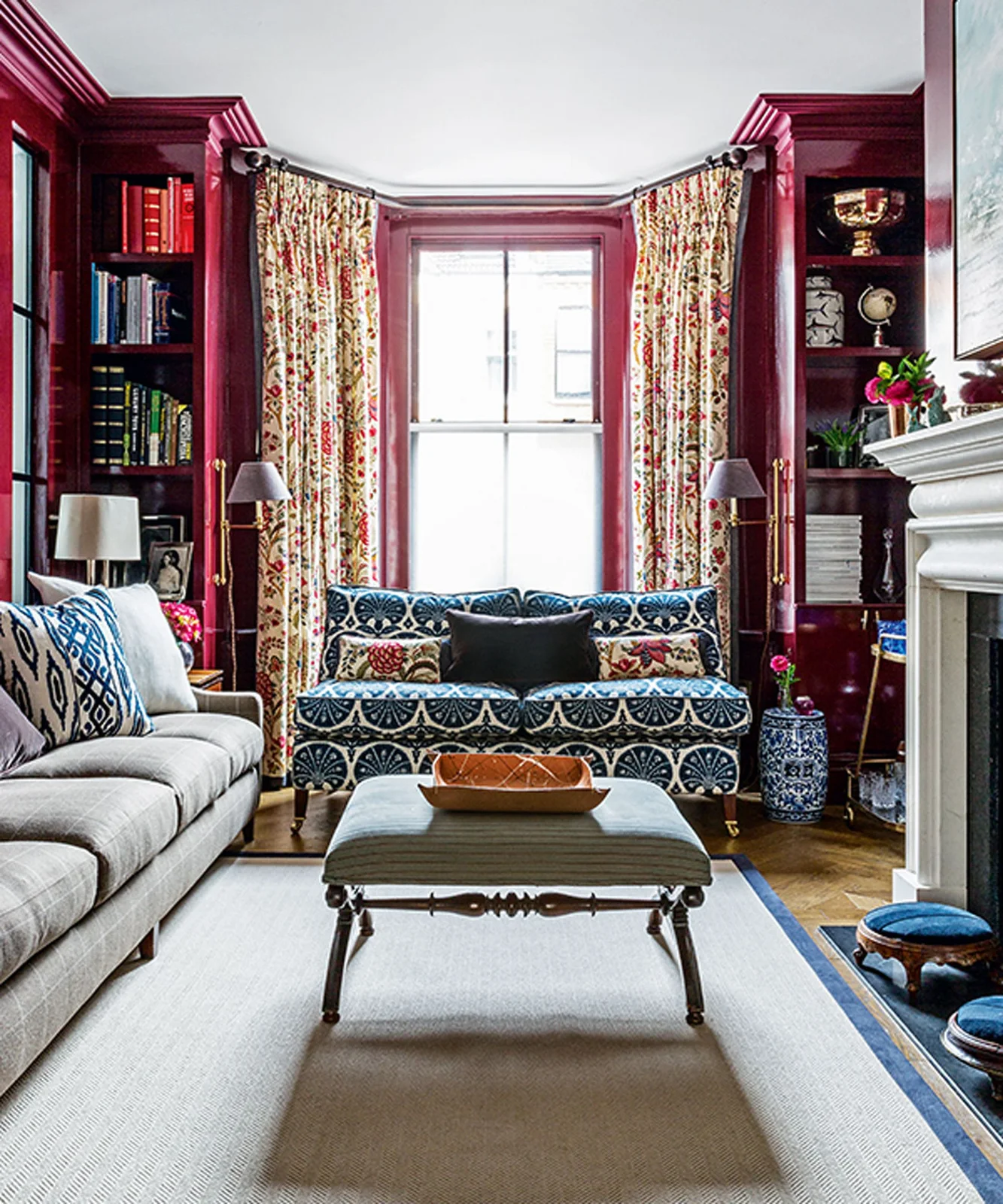 Red bookcases frame a window beside patterned curtains, a blue-and-white sofa and a central upholstered ottoman