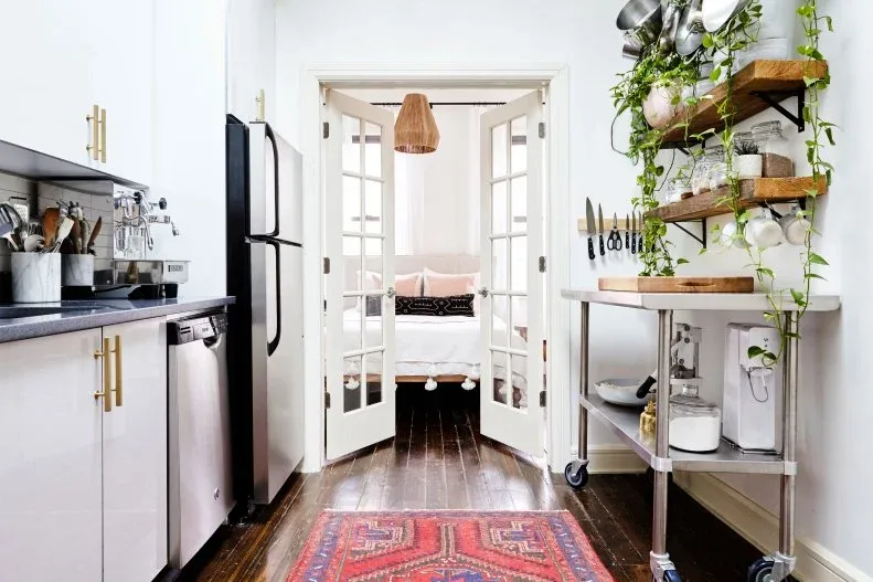White galley kitchen with trailing plants on wood shelves, a metal cart and a red patterned runner.
