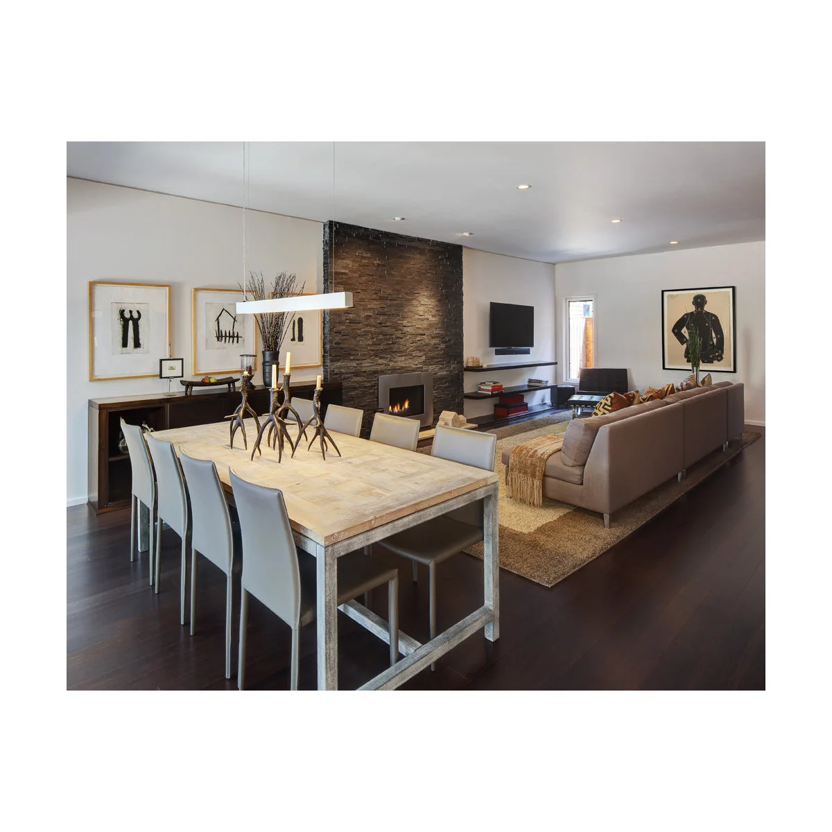 Long living dining room with a pale rectangular table, slim chairs and a brown sofa facing a fireplace and television beyond.