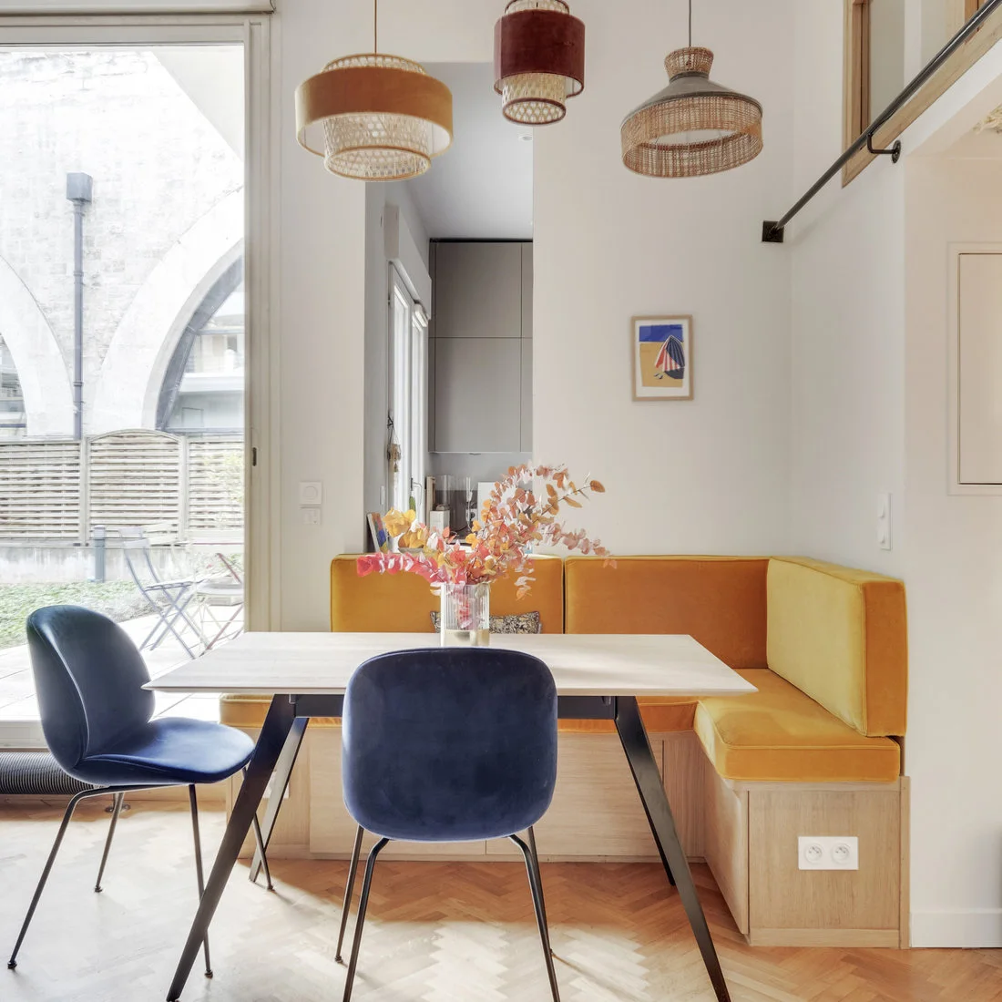 A mustard corner bench and navy chairs surround a pale dining table beneath three different woven pendant shades.