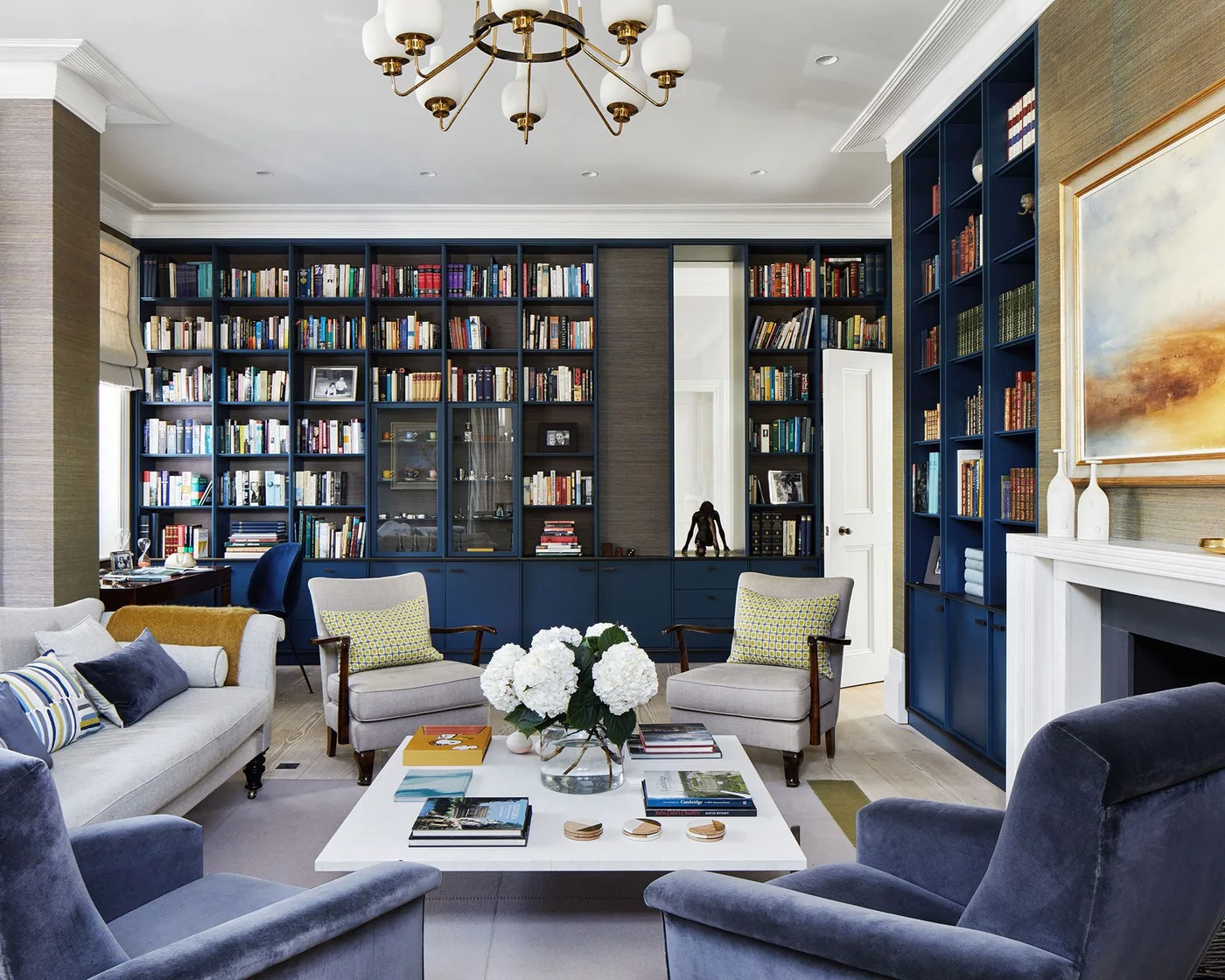 Dark blue bookcases line a sitting room around pale sofas, upholstered chairs, a white coffee table and a fireplace