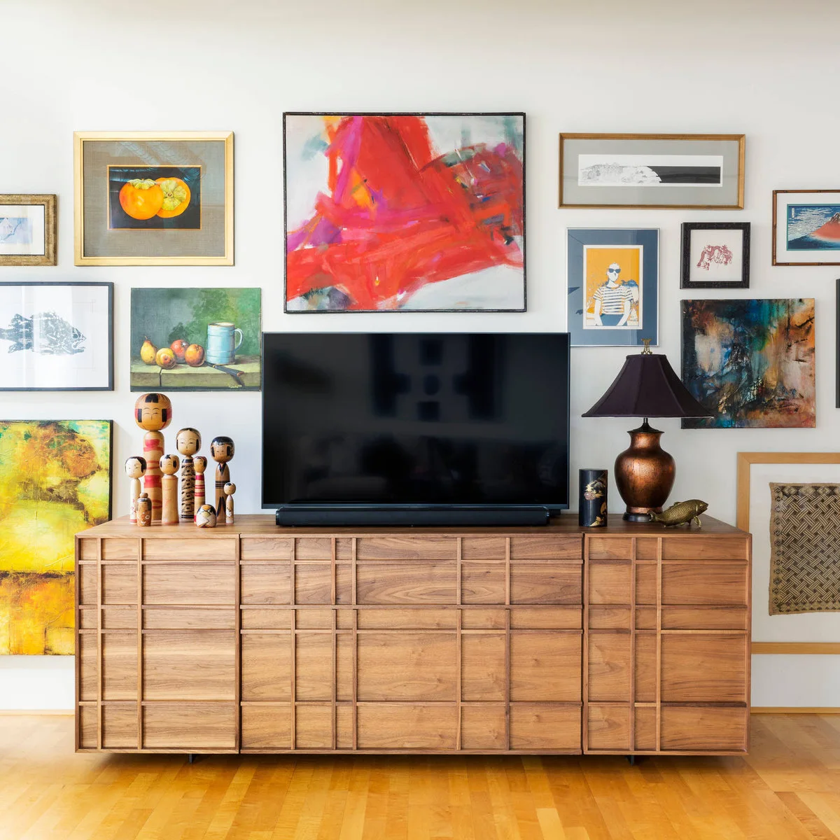 Red abstract painting above a television surrounded by varied artworks over a wide wood cabinet.
