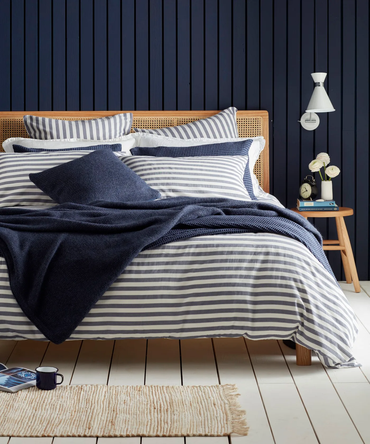 Navy vertical wall paneling behind a cane headboard with striped blue-and-white bedding and a white wall light.