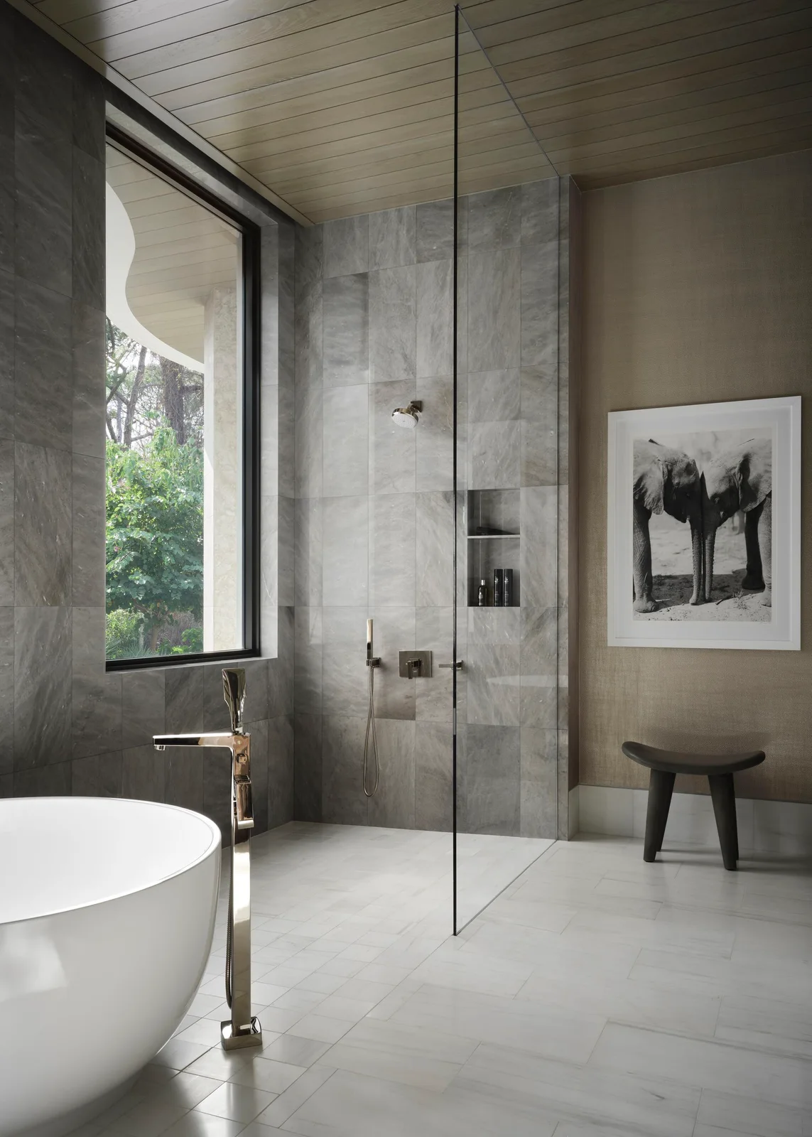 A clear shower screen separates gray large-format wall tiles from a white bath, black stool and pale floor beside a tall window.
