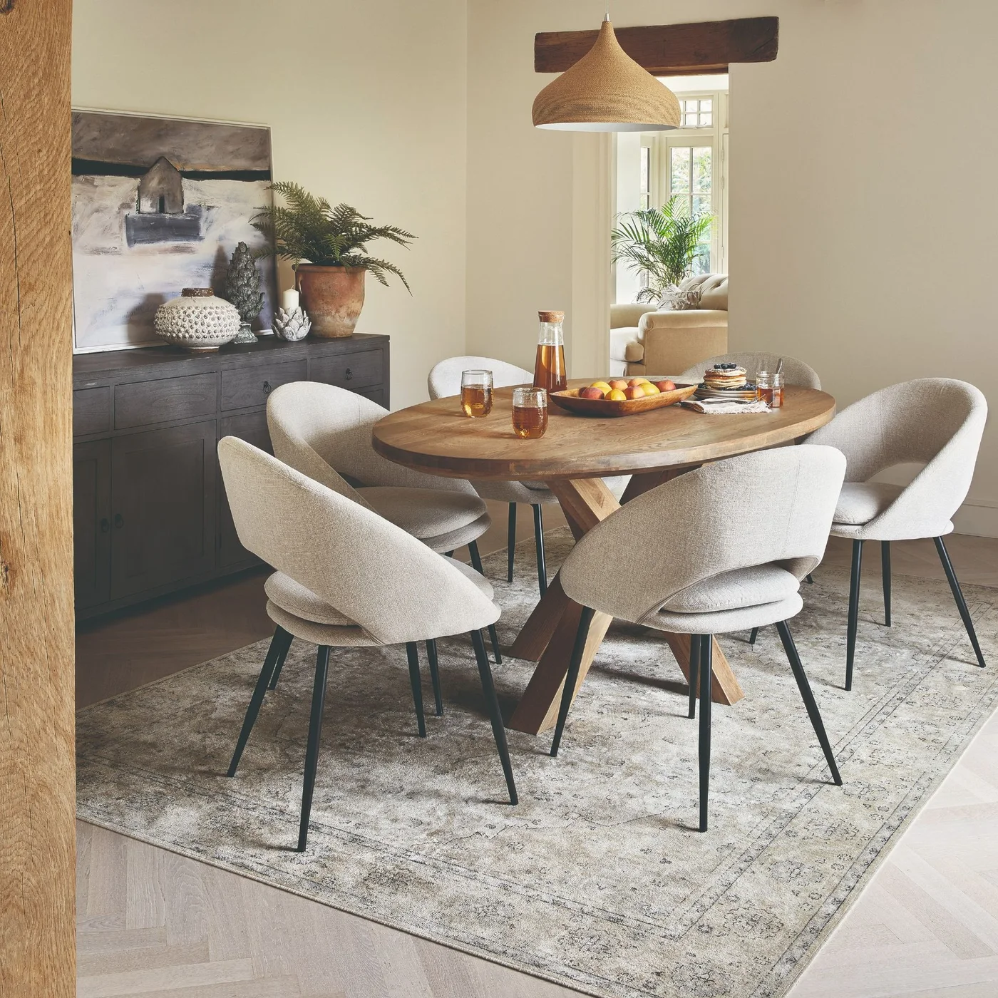 An oval wood dining table and pale upholstered chairs stand near a dark sideboard beneath a woven pendant in a neutral room.