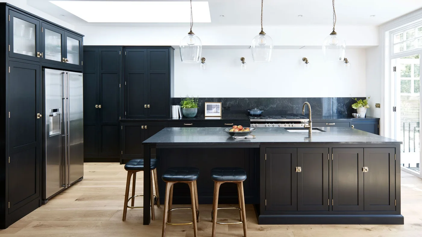 A dark kitchen island and tall cabinets stand beneath white walls, a skylight and clear pendant lights beside stools and a broad window