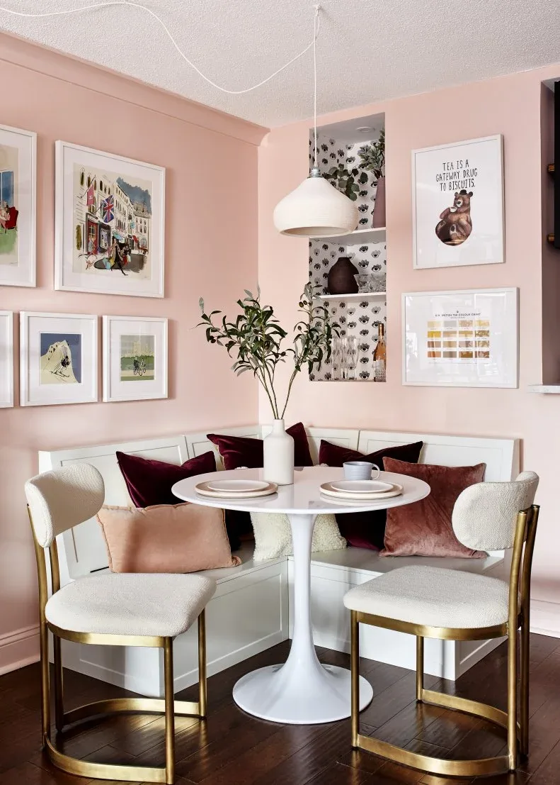 Pale corner banquette with burgundy and blush pillows against pink walls, with framed pictures, a round white table, and pale chairs with gold-colored legs.