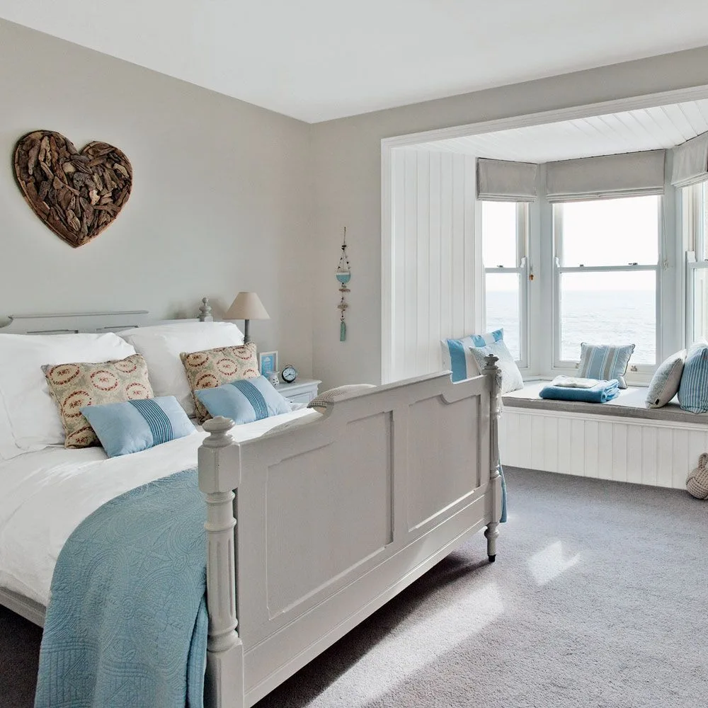 Pale gray bedroom with a white wooden bed, blue accents, a paneled bay-window seat and soft window blinds.