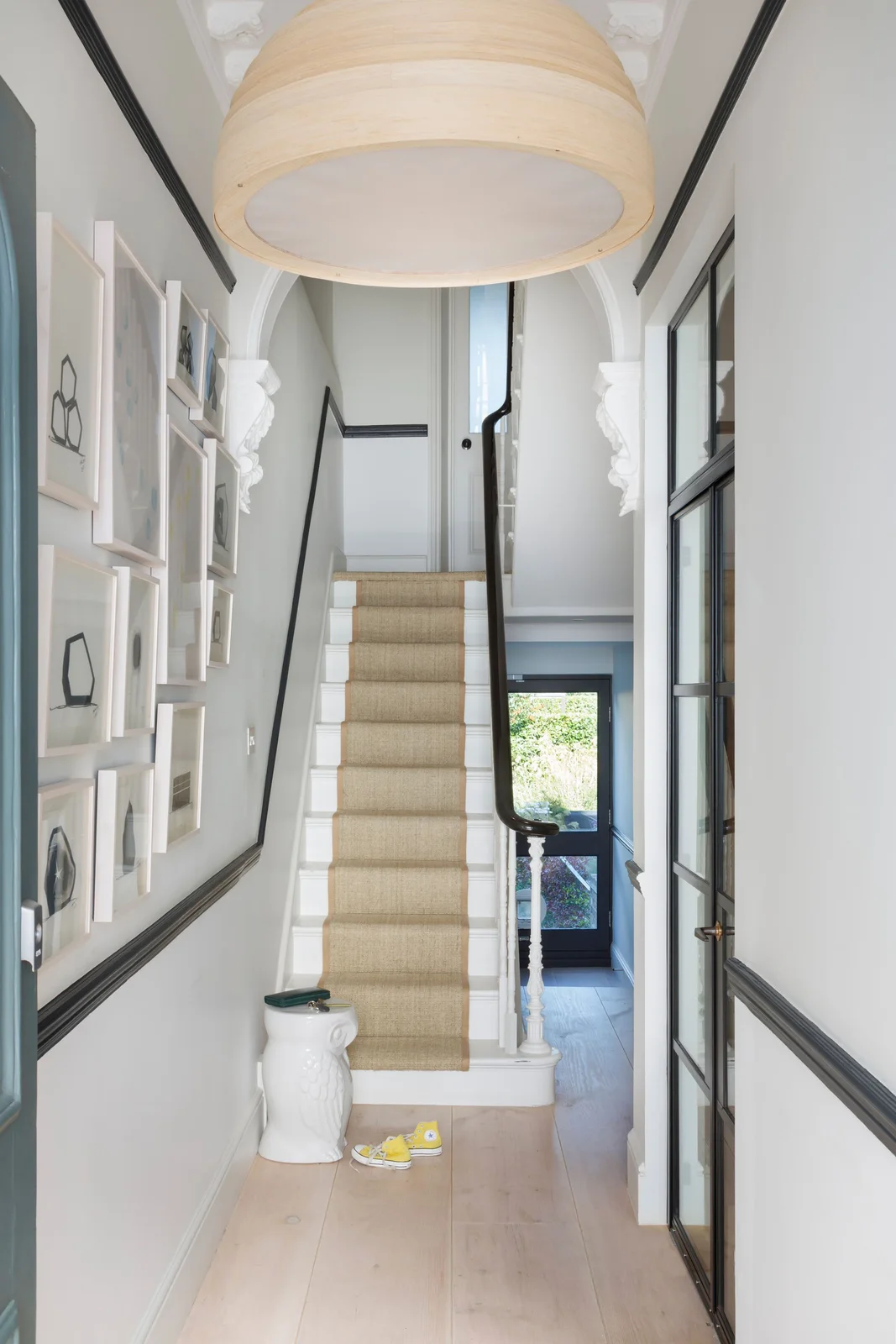 A narrow pale hallway has dark upper and lower trim lines, white-framed pictures, a pale stair runner and a dark-framed glazed opening