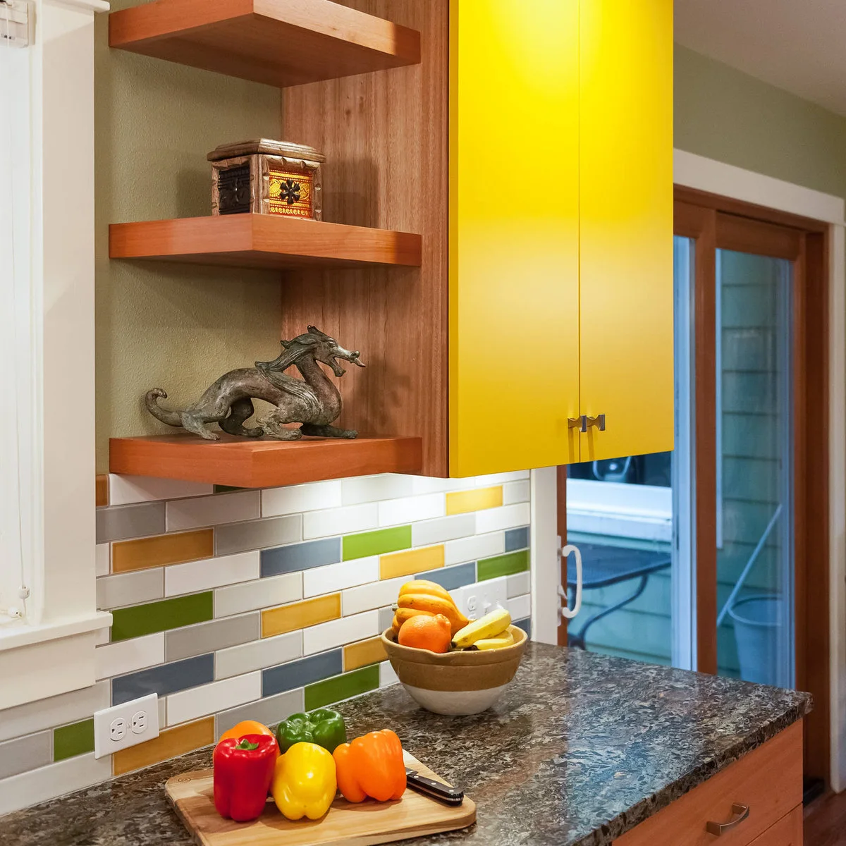 Yellow upper cupboard beside wood-colored shelves and a backsplash with yellow, green and gray tiles