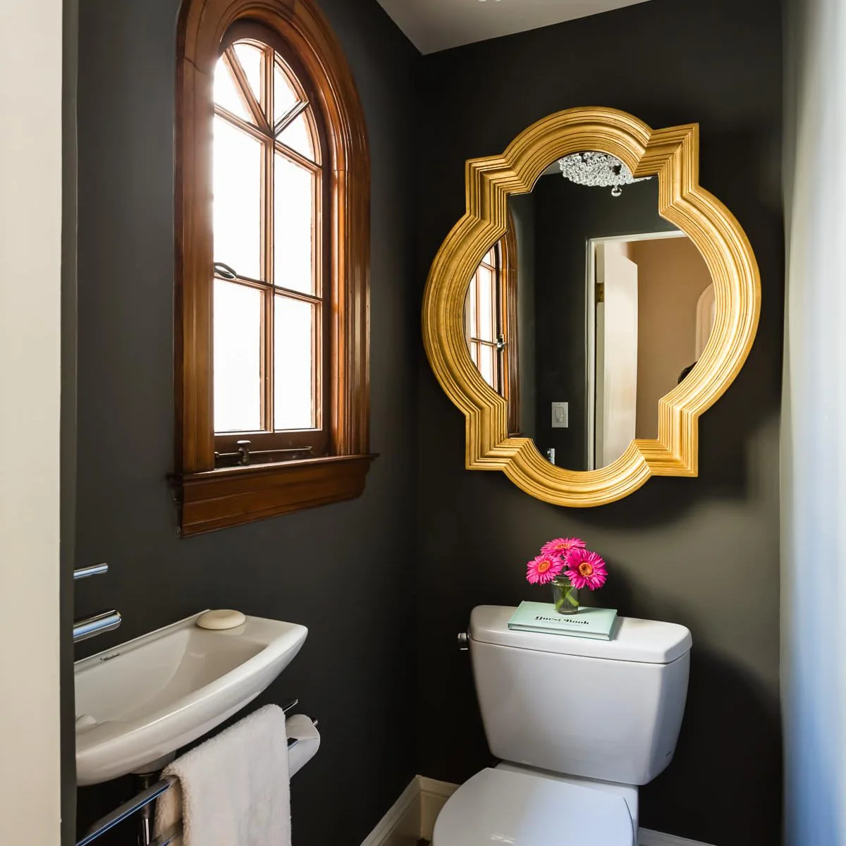 Gold-colored shaped mirror above a toilet beside an arched powder-room window