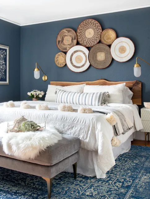 Round woven wall pieces form a cluster above a wood headboard and pale bedding against deep blue walls beside simple wall lights and a gray bench