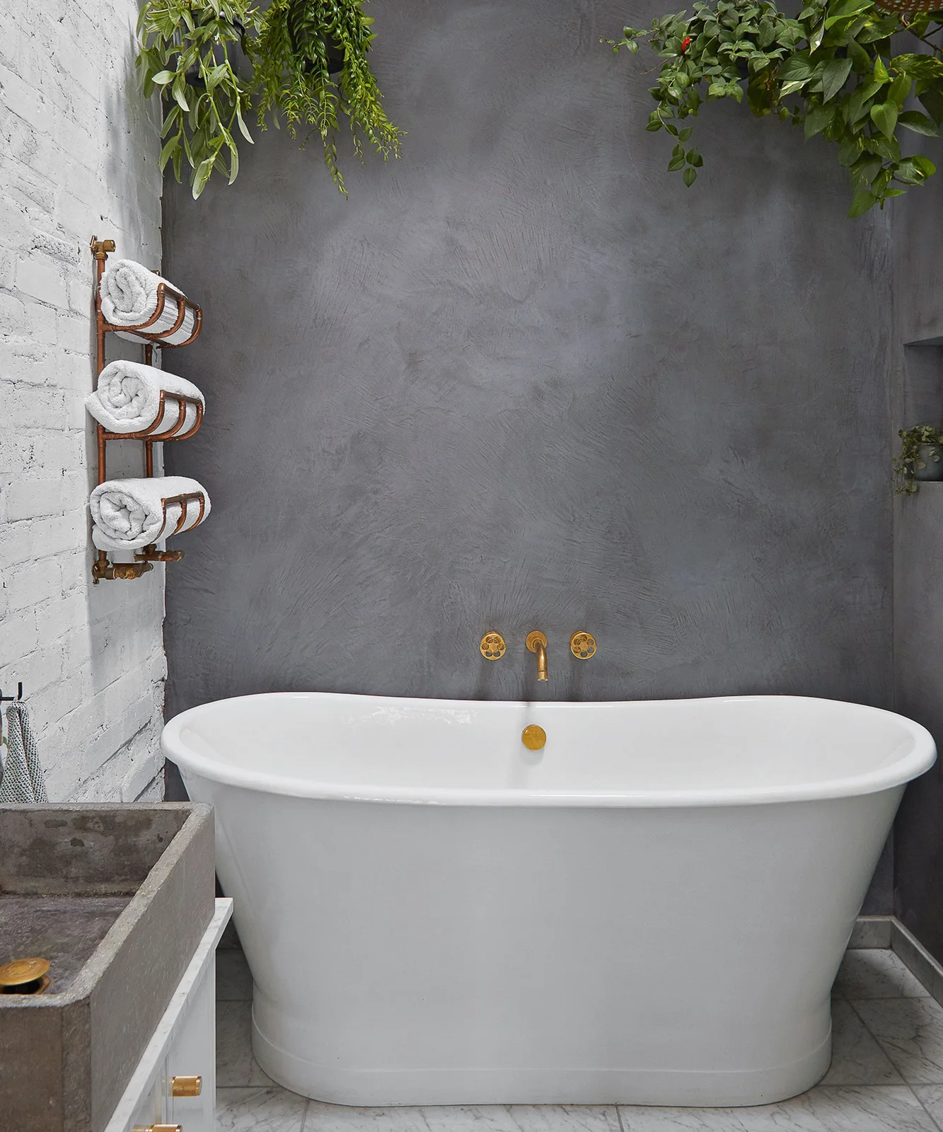 A white oval freestanding bath stands beneath warm-toned wall taps against a textured gray wall beside rolled towels, white brickwork and hanging plants