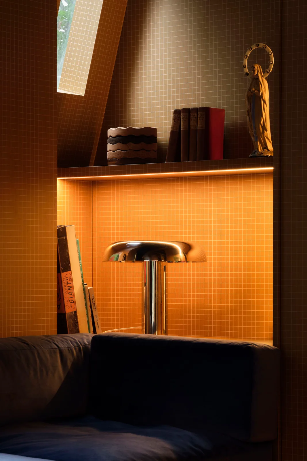 A navy upholstered bench sits beside warm tiled walls with an illuminated shelf of books, small objects and a reflective mushroom-shaped lamp