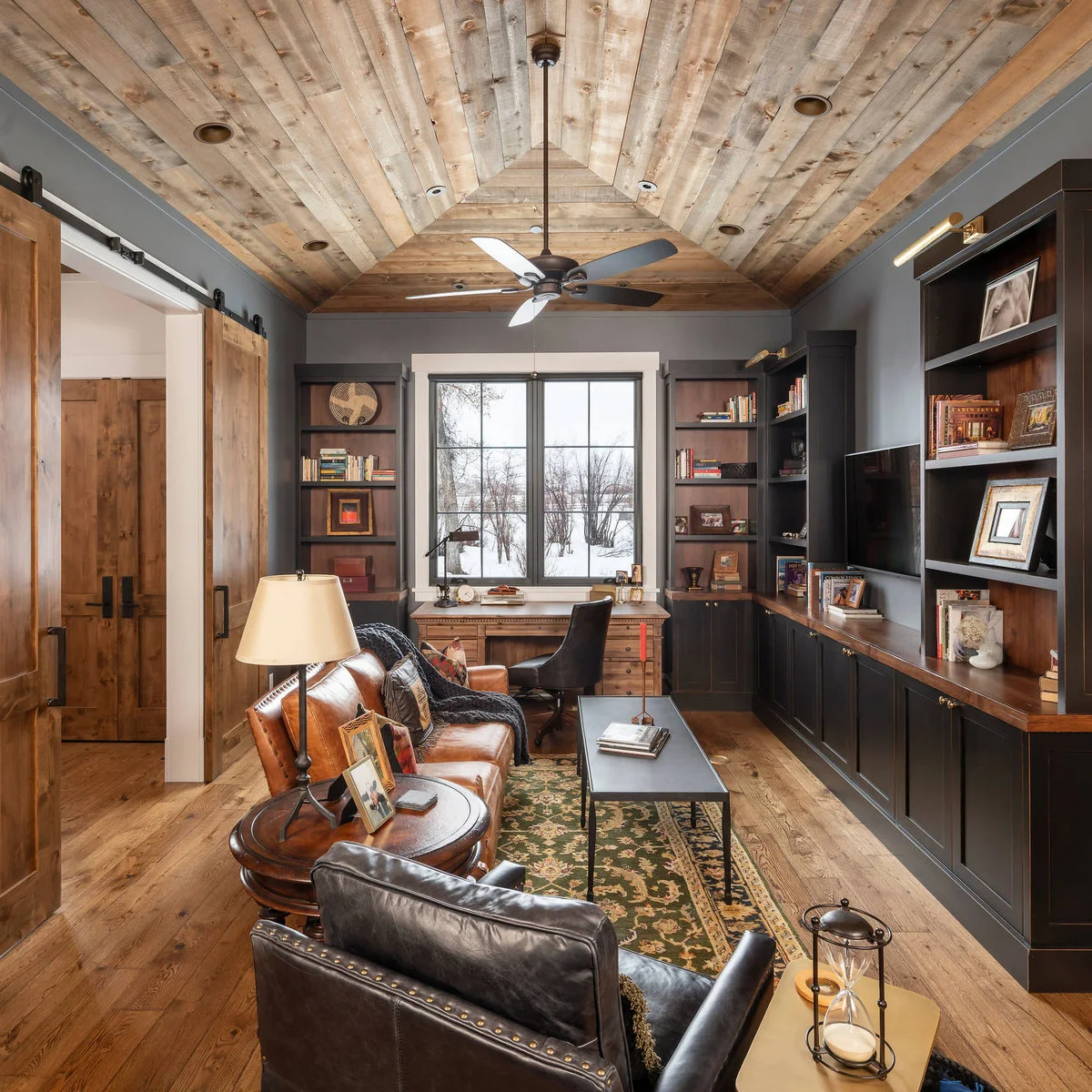 Study with pale wood ceiling, dark built-in cabinetry, wood desk at the window and brown sofa