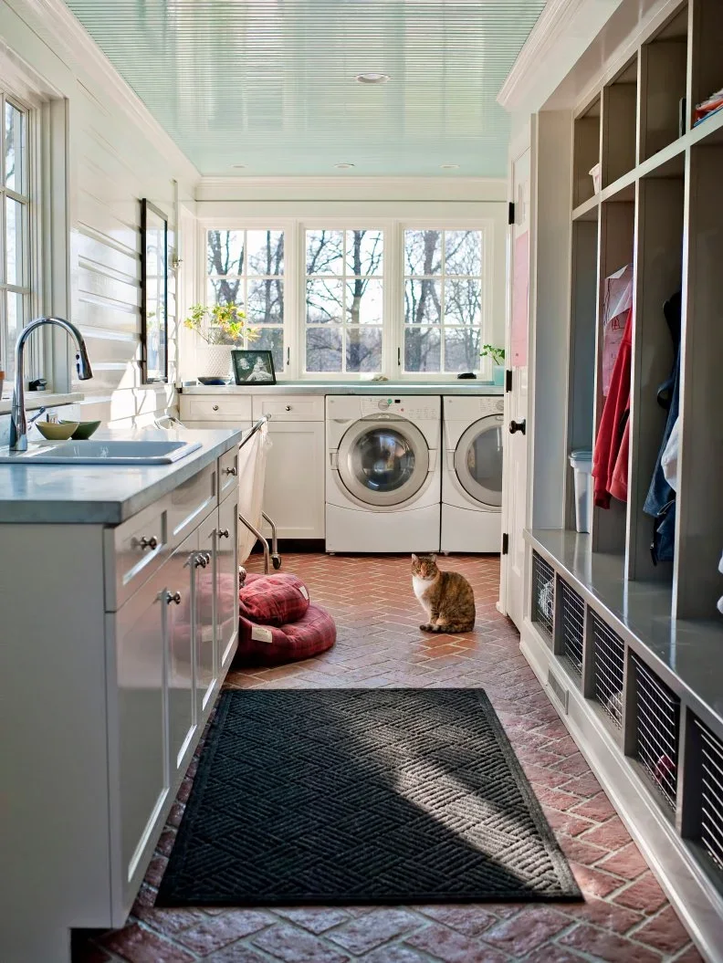 A narrow combined mudroom and laundry has white sink cabinets opposite open coat bays, with laundry machines below the far window.