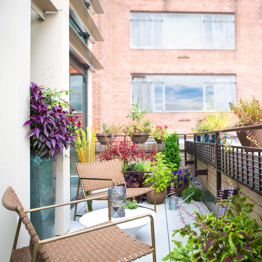Two woven chairs and a small white table sit among colorful container plants on a narrow balcony