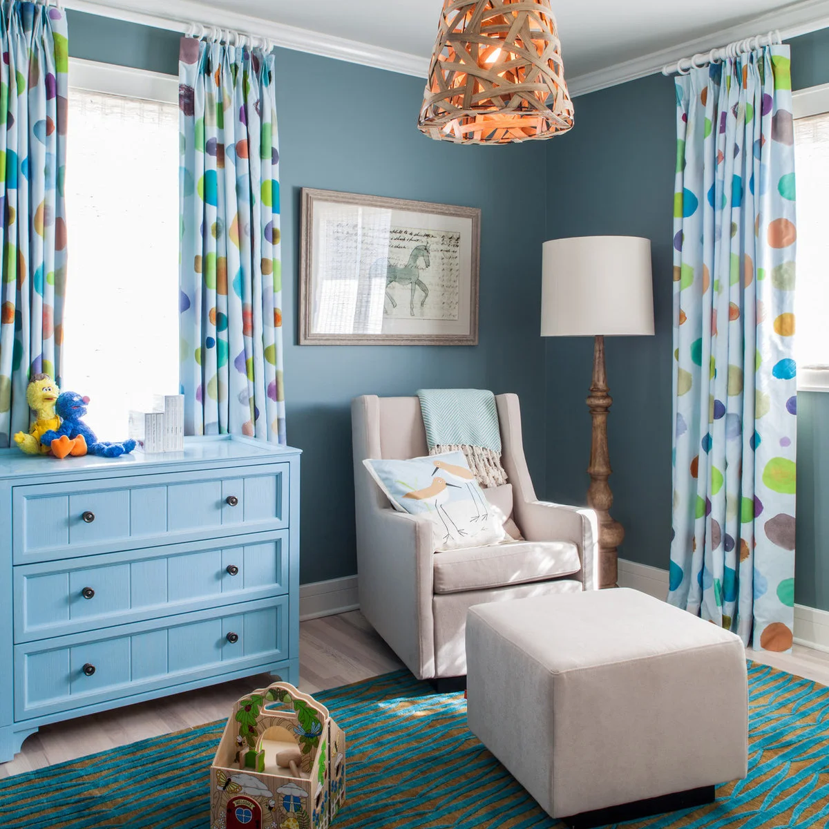 Blue nursery seating corner with multicolored curtains, a blue dresser and pale upholstered chair
