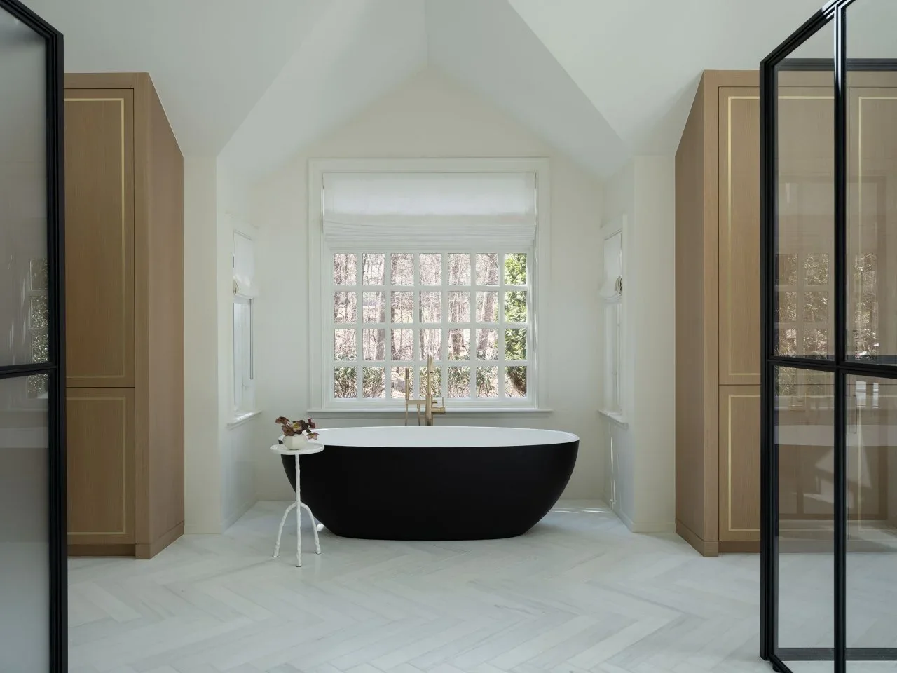 A black tub sits below a white window between two tall pale timber cupboards and black-framed glass doors.