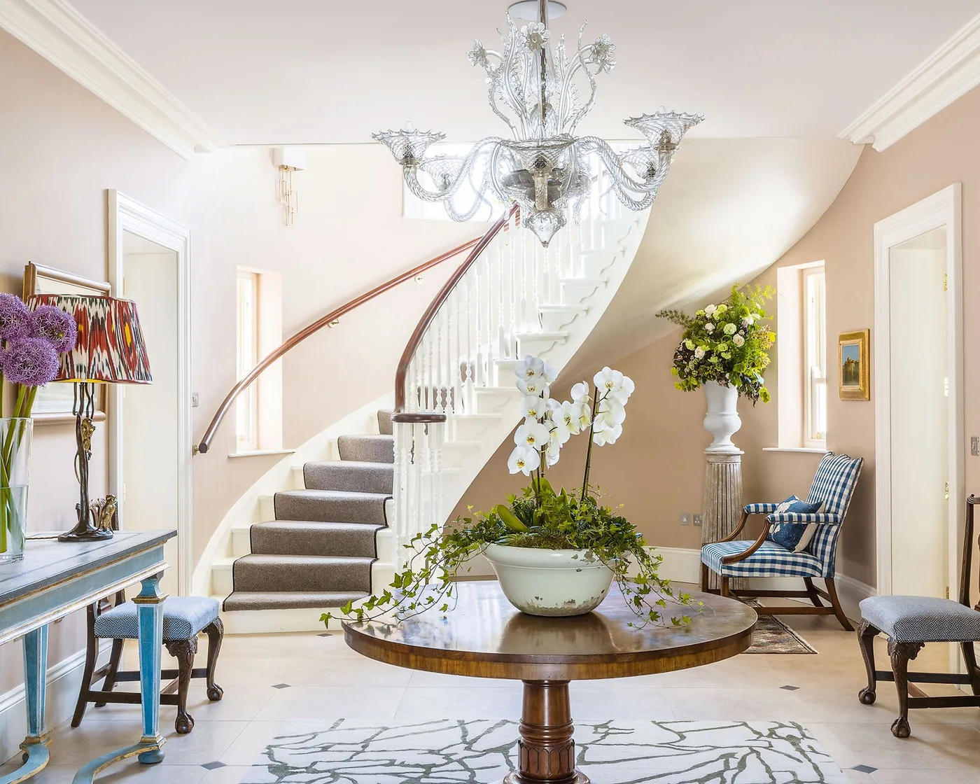 Clear curved-arm chandelier above round wood entry table, pale walls, gray stair runner, blue checked chair and painted console