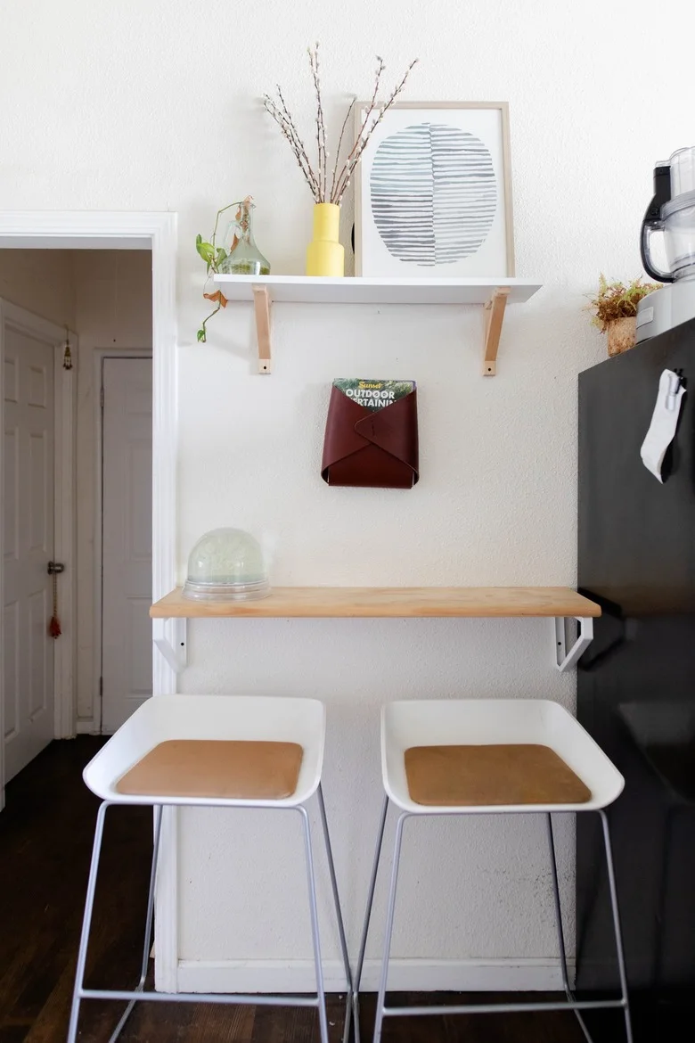 A narrow wood breakfast counter is fixed to a white wall beside a dark refrigerator, with two white stools, a shallow shelf and a small framed print.