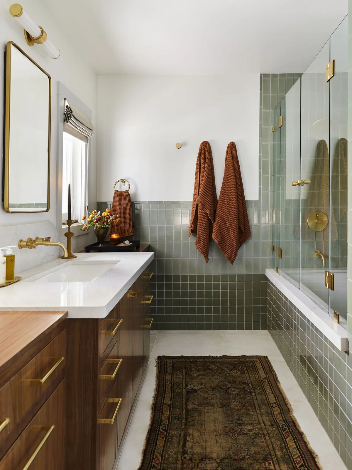A bathroom combines deep olive square tiles with softer sage rectangular tiles, a wood vanity, brass hardware and orange towels.