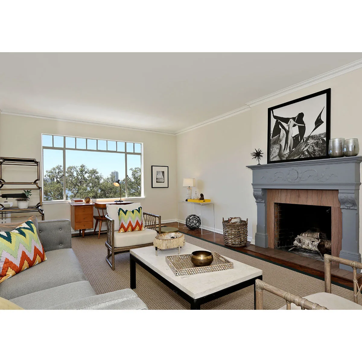 A wood desk beneath a broad window at the edge of a living room with gray seating and a fireplace.