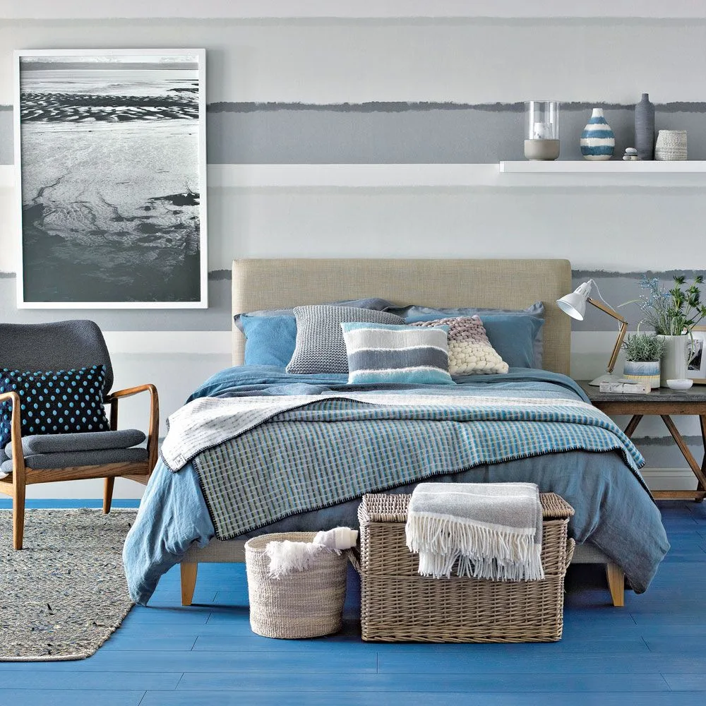 Bedroom with blue painted floorboards, pale upholstered bed, blue bedding, gray-and-white horizontal wall bands and storage baskets.