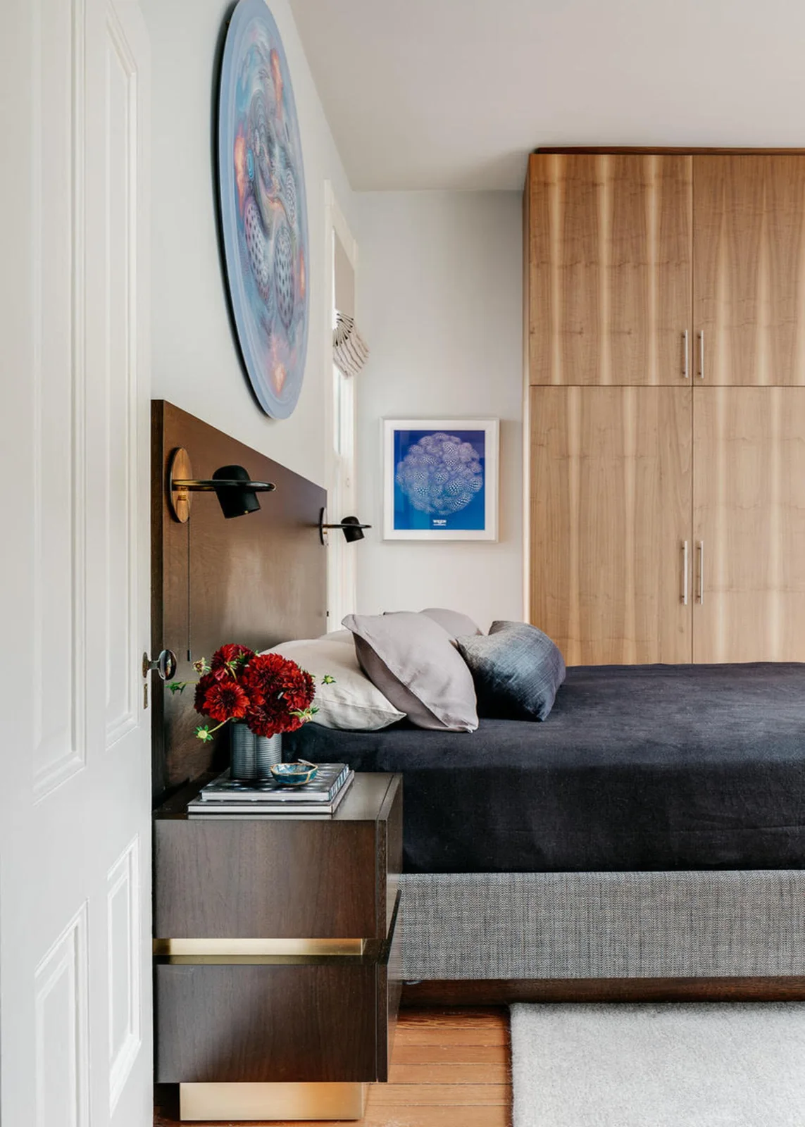 A wood bedroom headboard and tall wardrobe sit beside dark bedding, a wood bedside cabinet, small wall lights and colorful framed artwork