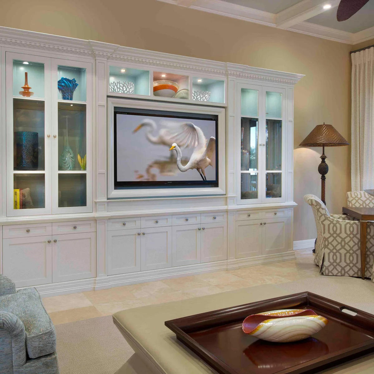 A white media wall has glass-front side cabinets, small illuminated display cubbies above the television and plain lower cupboards.
