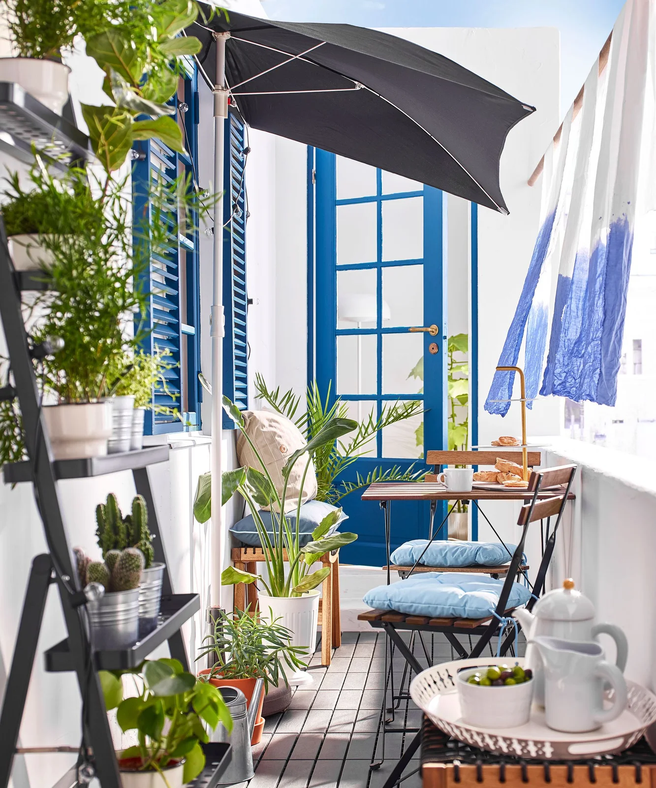 A white-and-blue curtain hangs beside a narrow balcony dining set beneath a dark umbrella