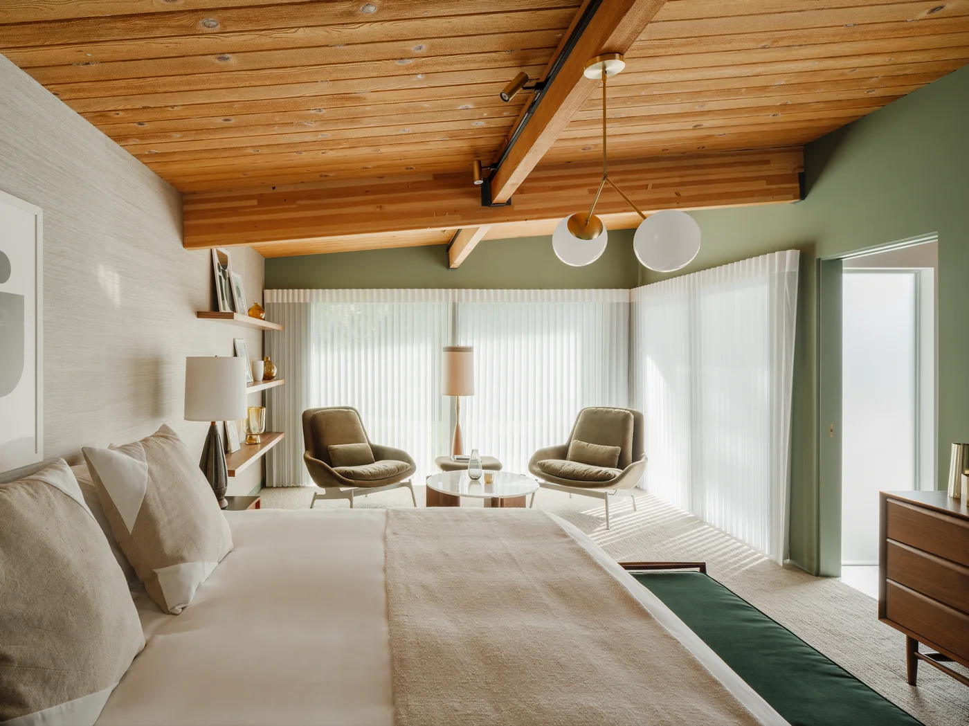 Muted sage walls frame broad windows below a timber ceiling in a bedroom with pale bedding and a green bench.