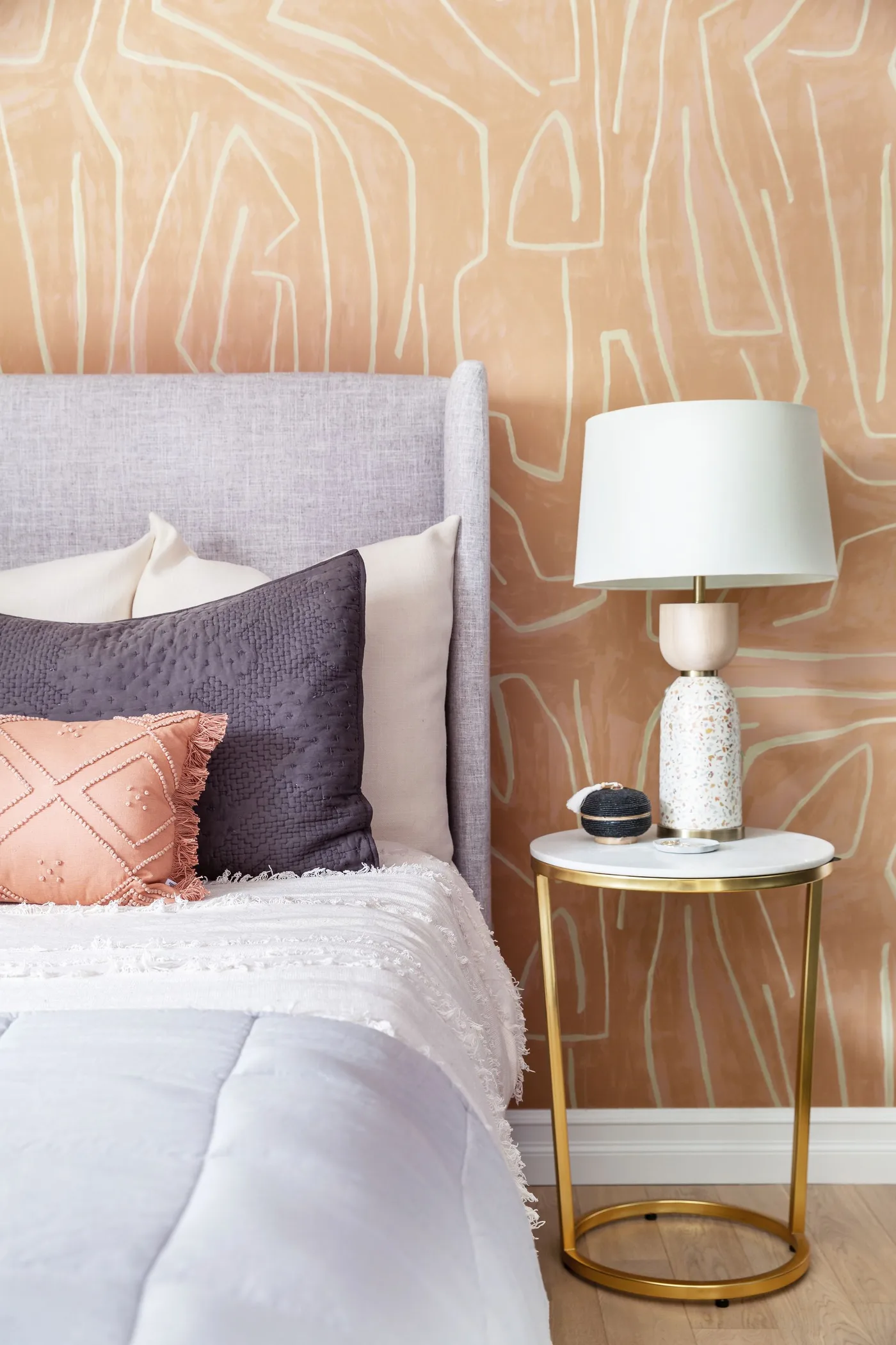 Bedroom with clay-colored abstract wallpaper, a gray headboard, peach and navy cushions and a round gold-colored bedside table