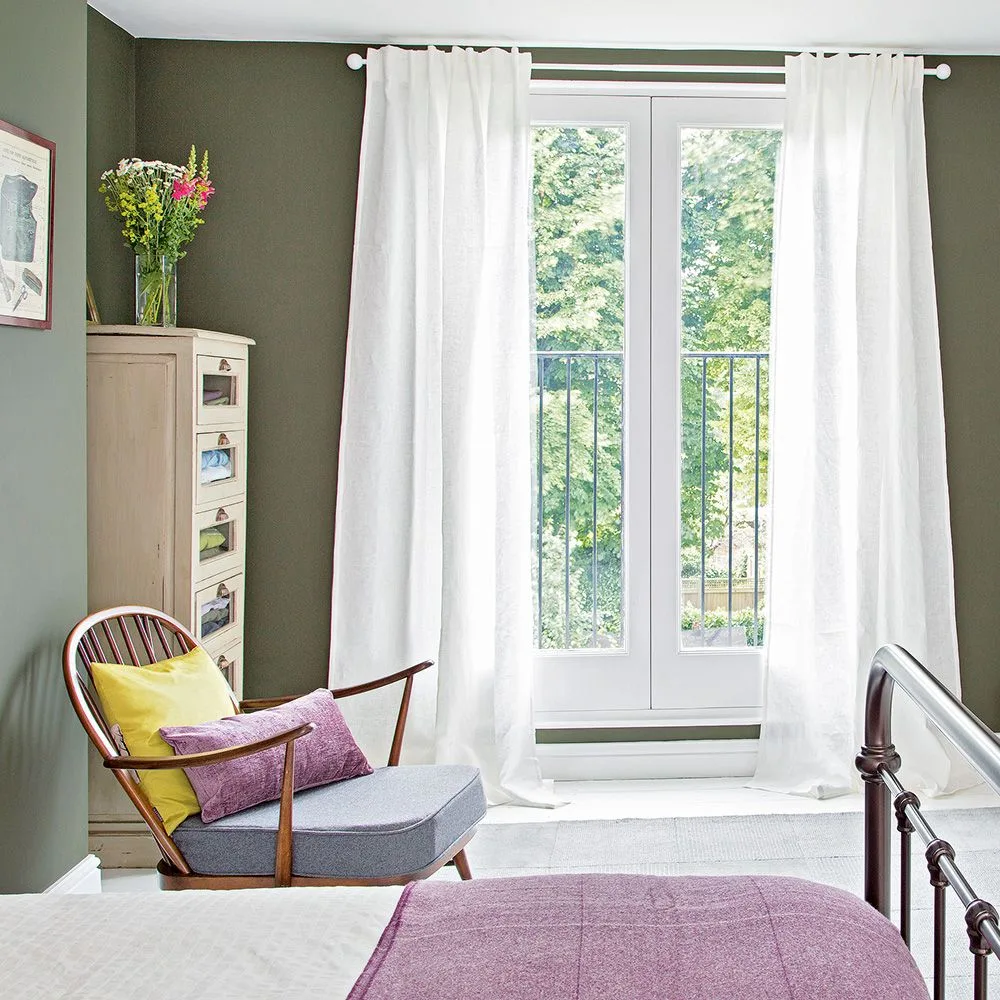 Olive bedroom walls around a tall window with long pale curtains, cream storage chest, wooden chair, colorful cushions, and pink bedcover.
