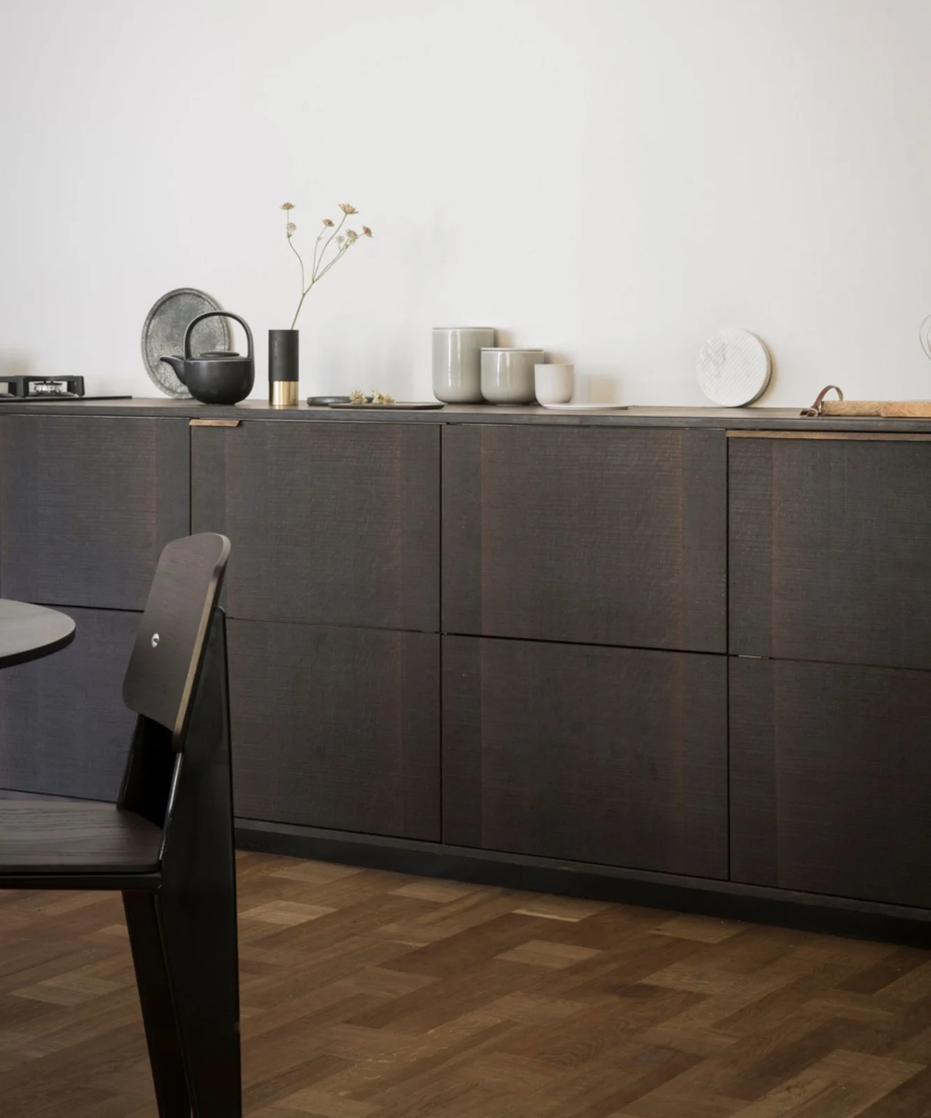 Dark wooden lower cabinets sit against a plain white wall beneath pale cups, a black kettle and a narrow vase beside a dining chair