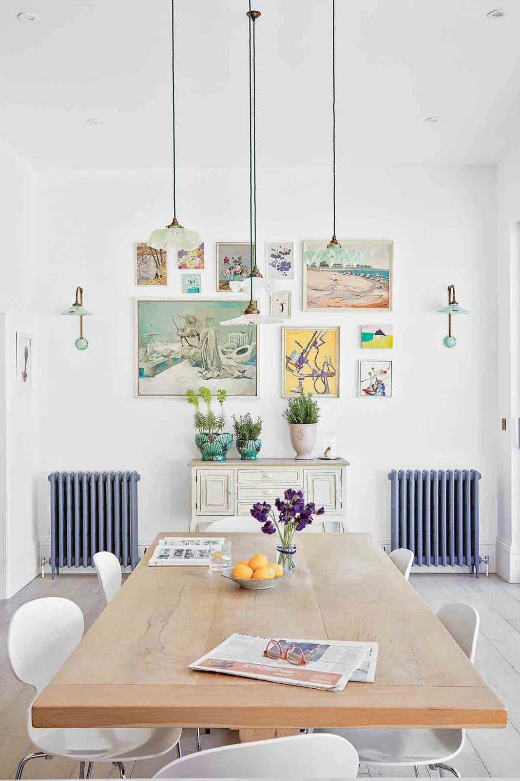 Pale molded dining chairs on slim metal bases surround a wood table beneath green pendant lights beside a gallery wall, light floorboards and blue radiators