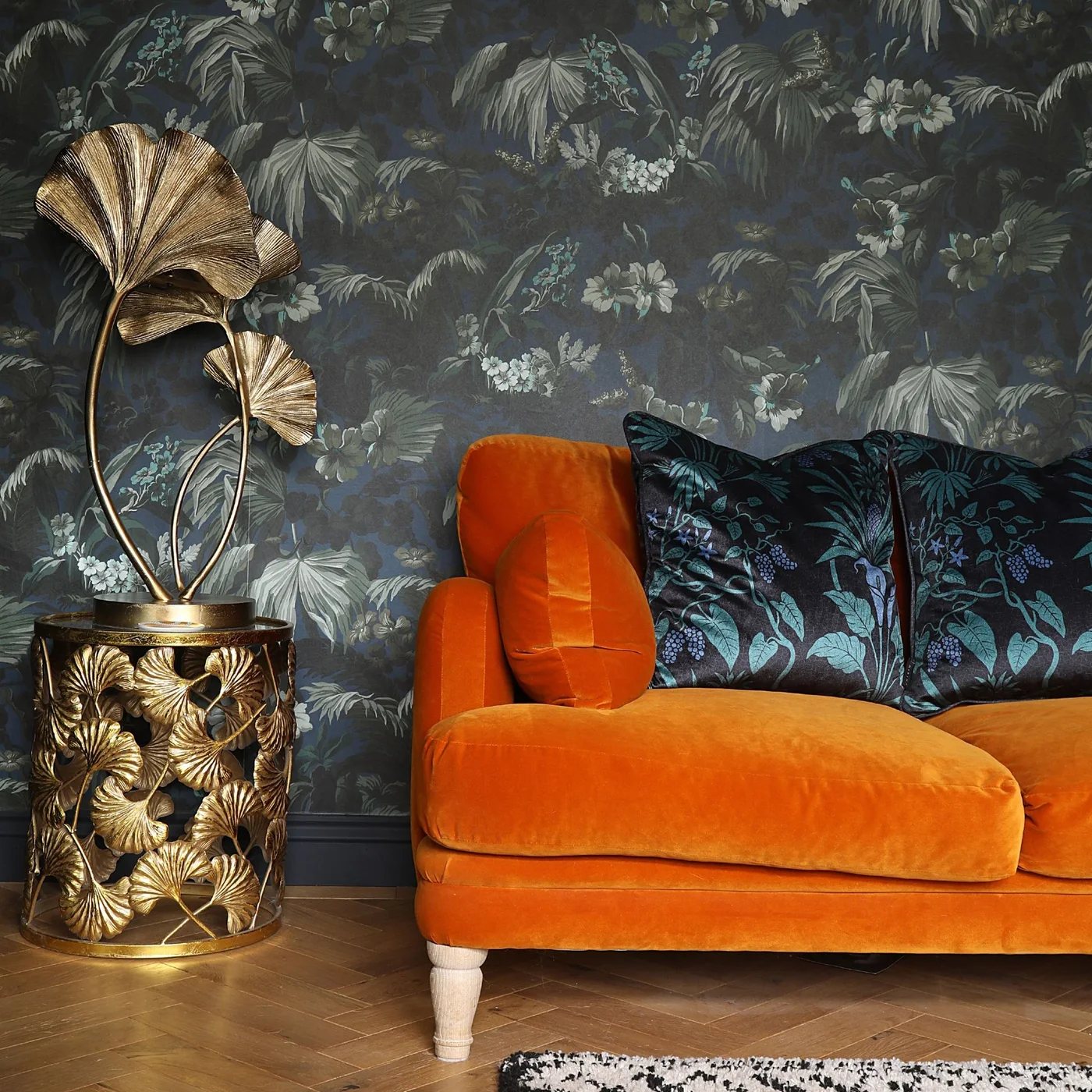 An orange sofa sits against dark botanical wallpaper beside a patterned warm-metal table and leaf-shaped lamp.