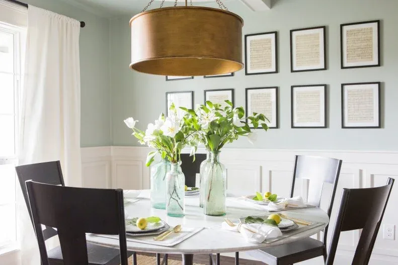 Eight framed pale pages in a two-by-four grid above white dining-room paneling, with a pale wooden table, dark chairs, a drum pendant, and flowers.