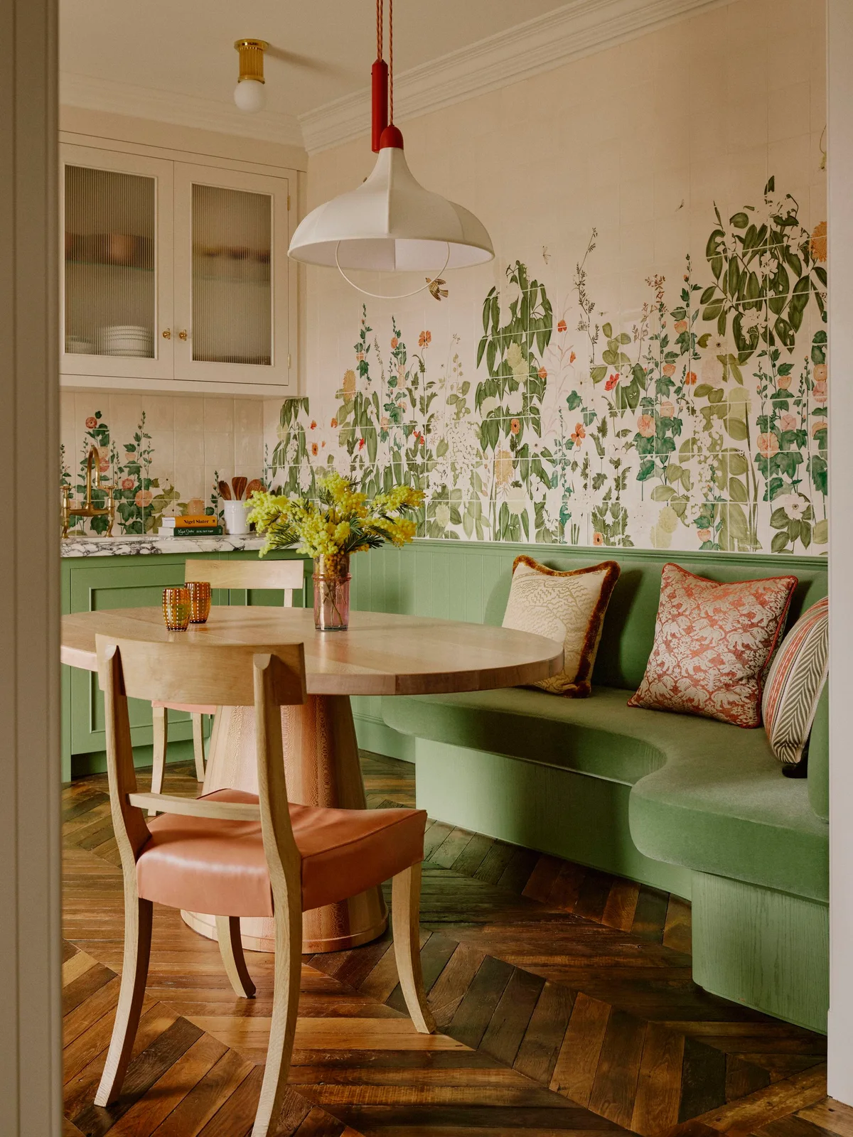 Leafy illustrated tiles surround a green curved kitchen bench with patterned cushions beside green cabinets and a wooden round table