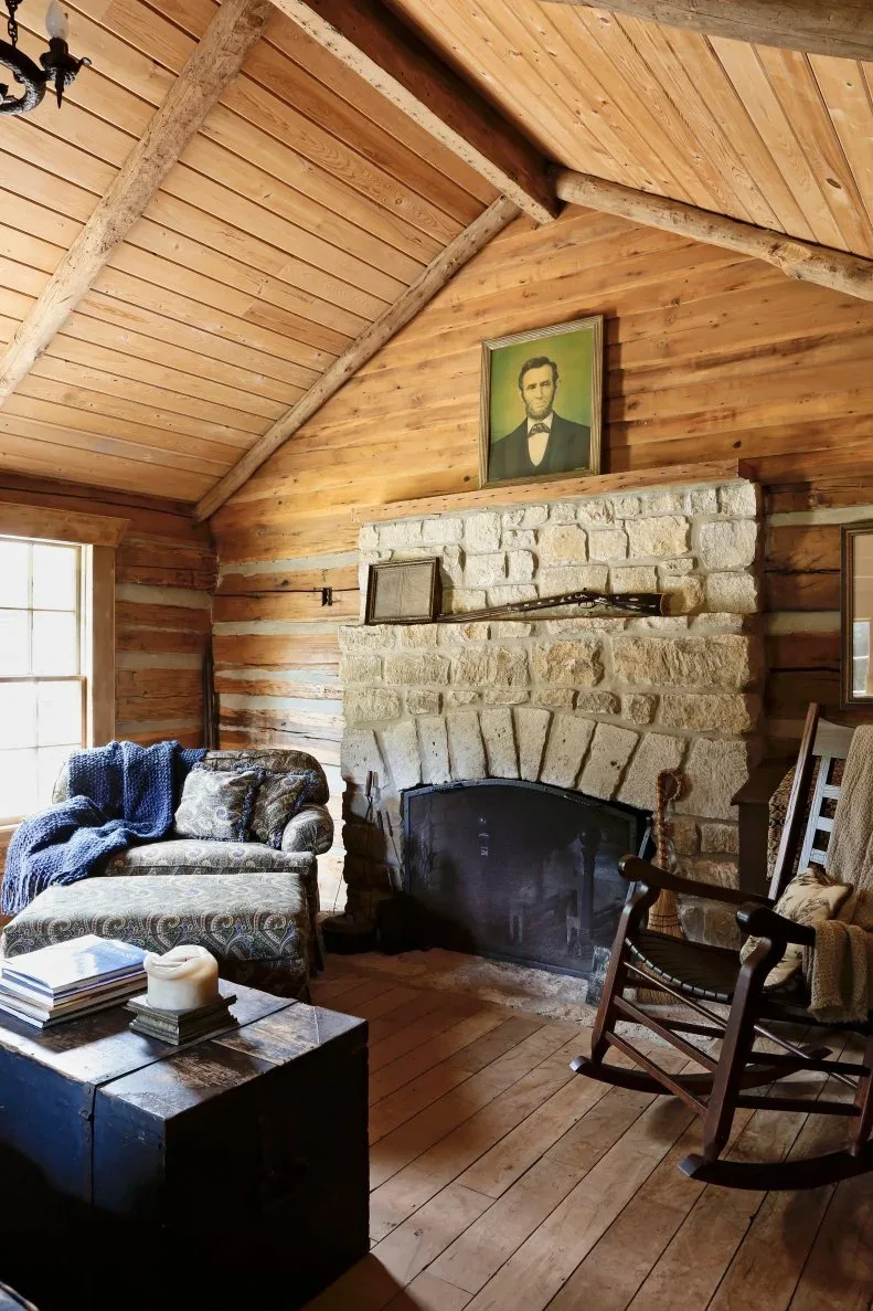 Wood-lined country room with stone fireplace, dark rocking chair, upholstered armchair, low trunk table and portrait above mantel