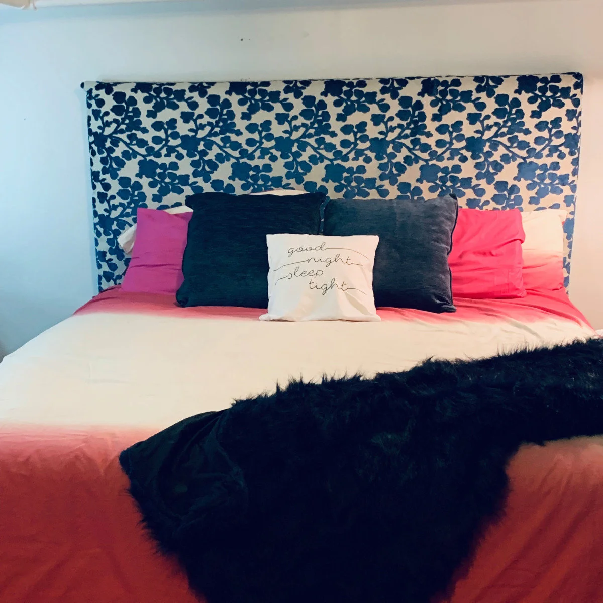College-room bed with a blue floral headboard panel, pink bedding, navy cushions and a dark throw